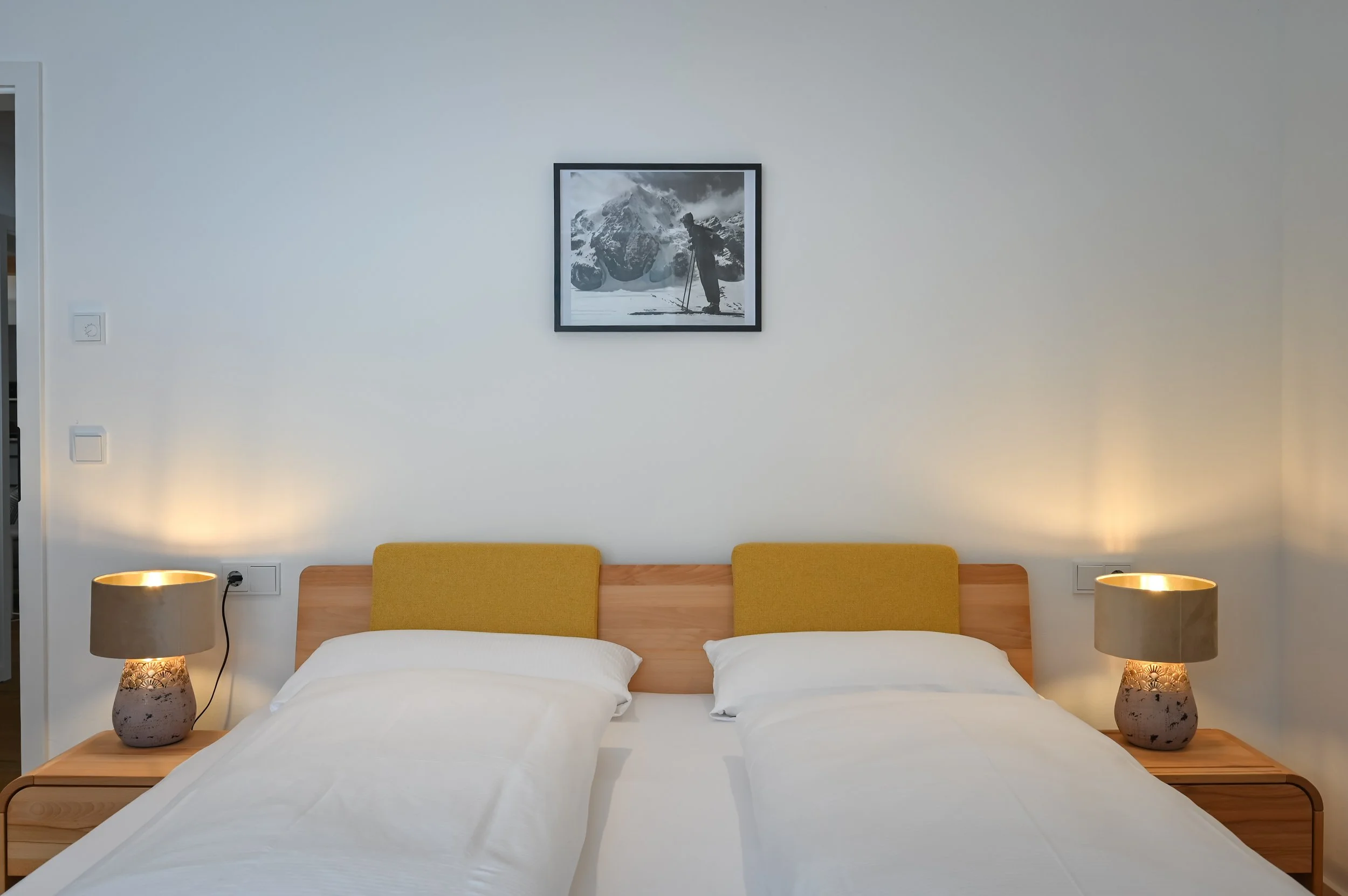 Schlafzimmer mit zwei Einzelbetten, Nachttische mit Lampen, Wandbild eines Berges, moderne Einrichtung, weißer Wand, warme Beleuchtung.