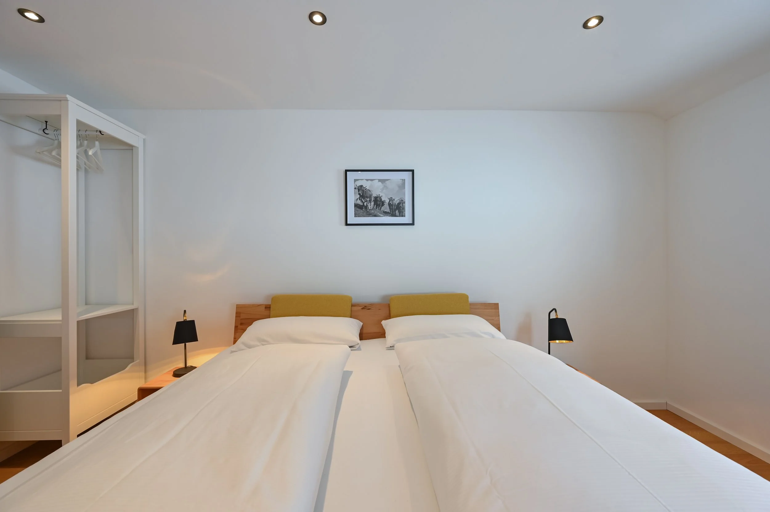 Schlafzimmer mit zwei getrennten Betten, zwei Nachttischen mit schwarzen Lampen, einer weißen Möbelstange auf der linken Seite, einem einfachen Rahmen mit schwarzem und weißen Foto an der weißen Wand, und Holzfußboden.