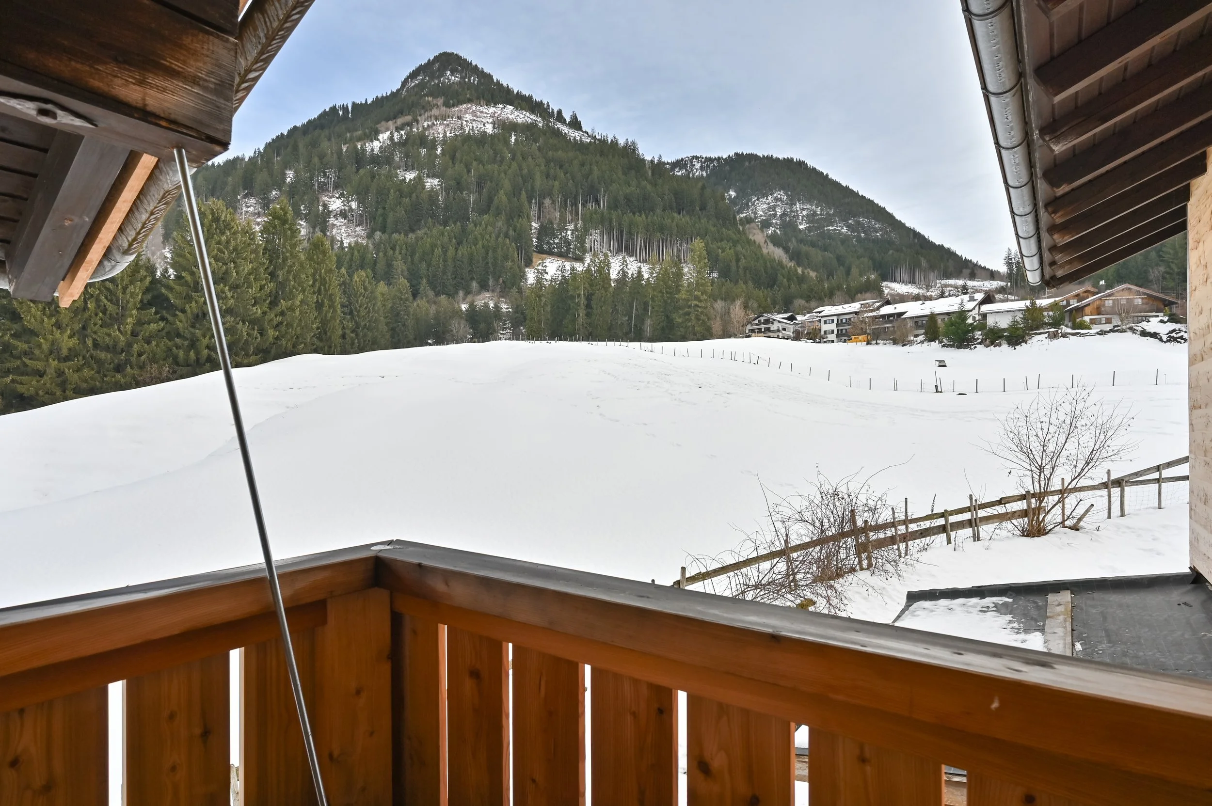 Blick von einem Balkon auf eine verschneite Landschaft mit Bäumen, Hügeln und einem Dorf im Hintergrund, auf einem winterlichen Tag.
