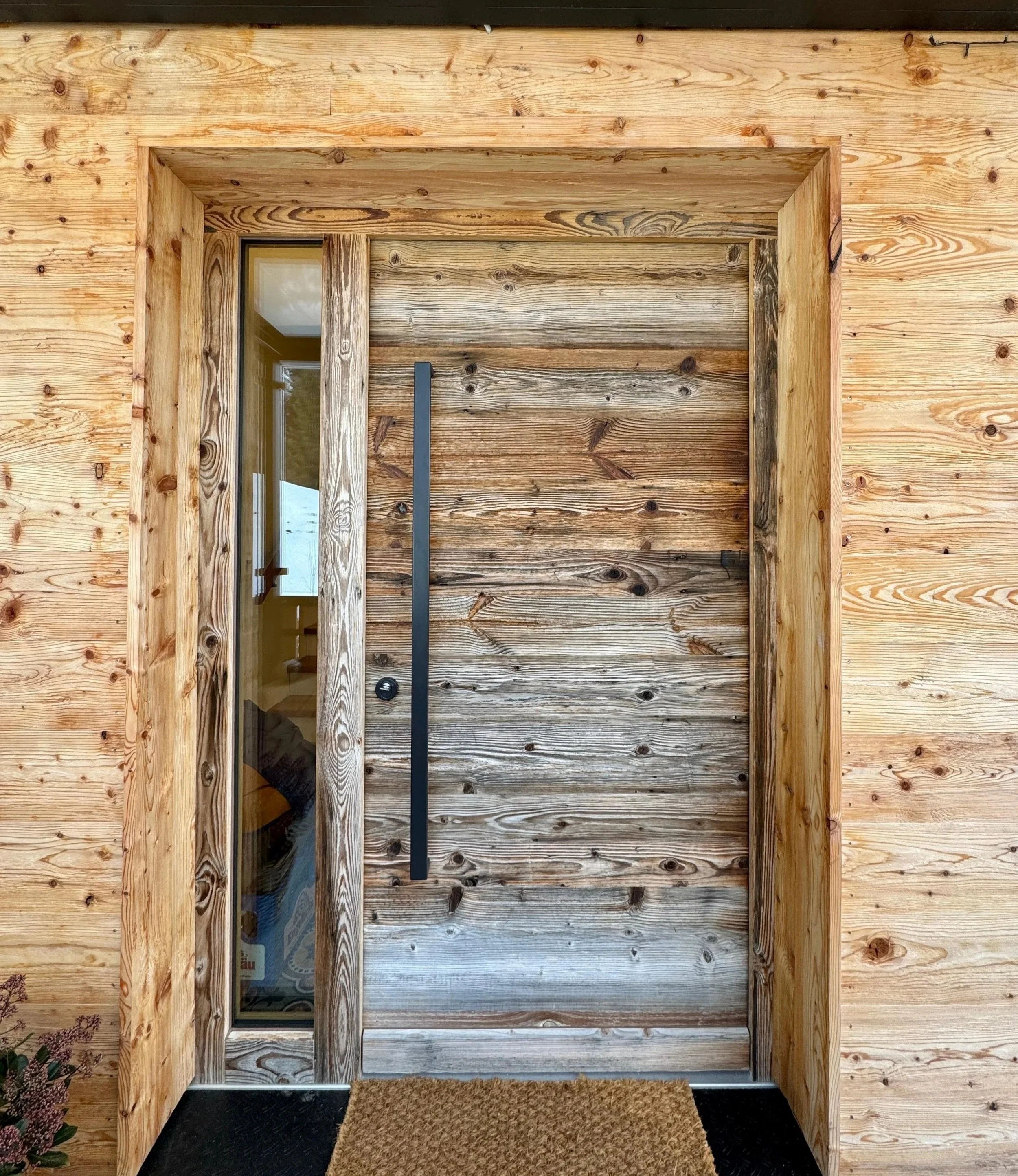 Holztür mit Glasfenster und schwarzem Griff vor einer Holzfassade, mit einem Türvorleger davor.