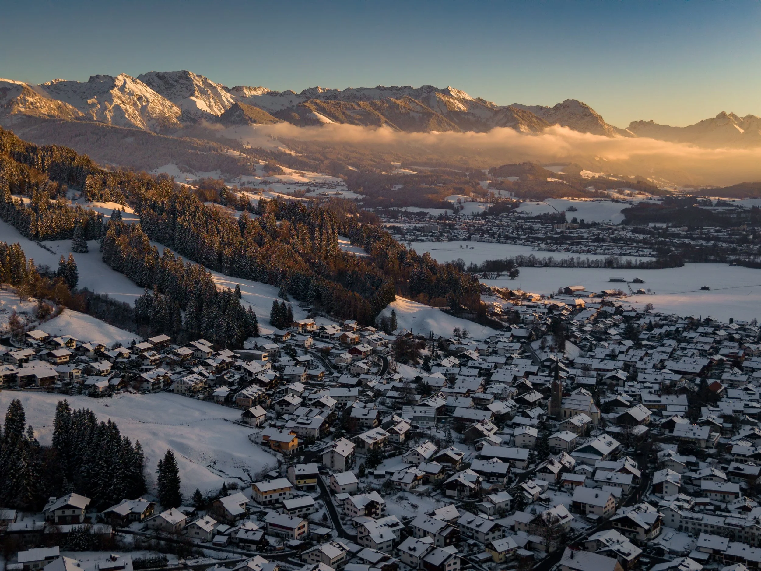 Eine verschneite alpine Landschaft mit einem Dorf, umgeben von Bergen und Wäldern bei Sonnenuntergang.