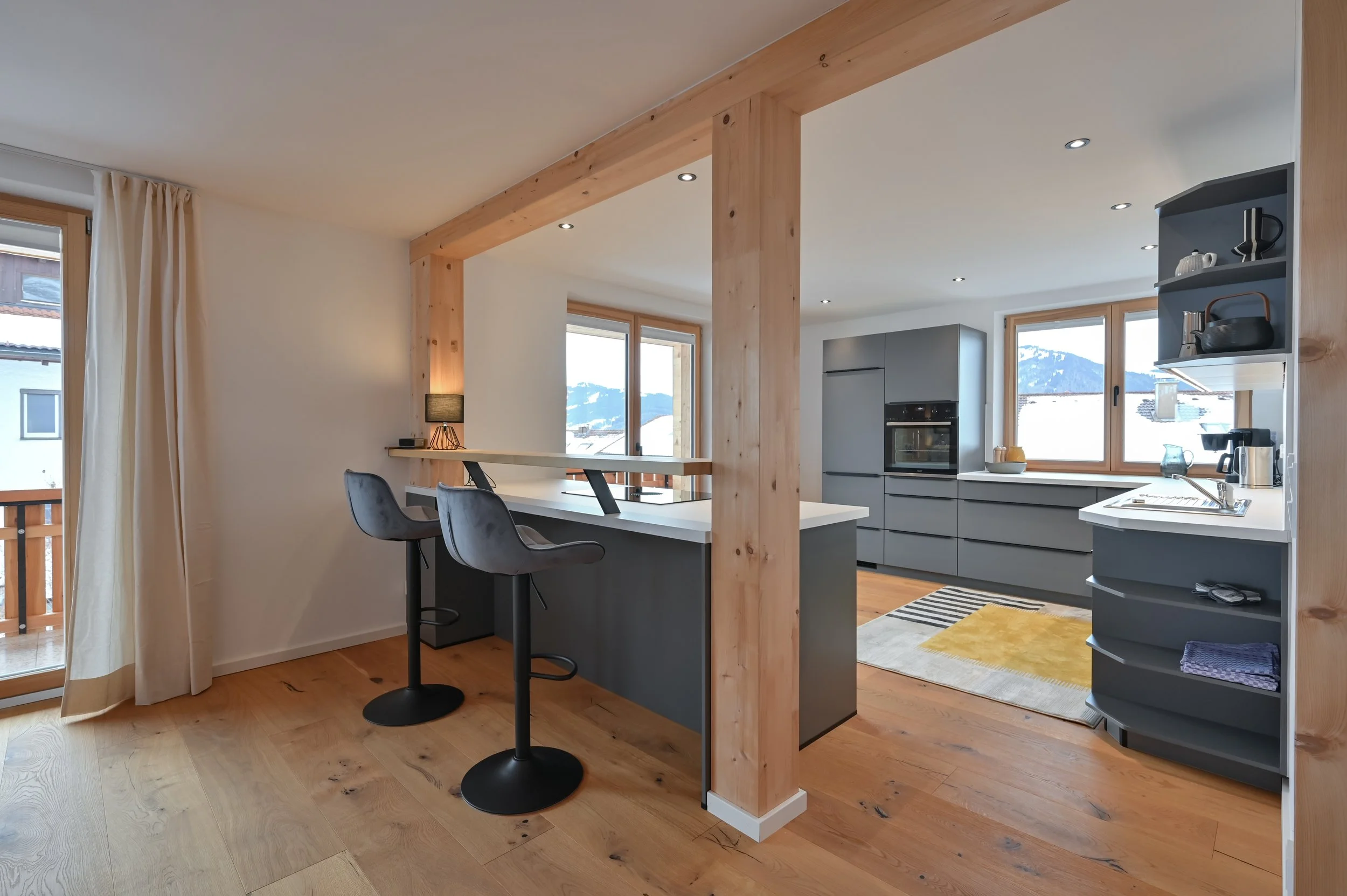 Moderne Küche mit grauen Schränken, Holzfußboden, Blick auf Fenster mit Bergblick, Esstheke mit zwei schwarzen Barhockern, offene Regale mit Geschirr