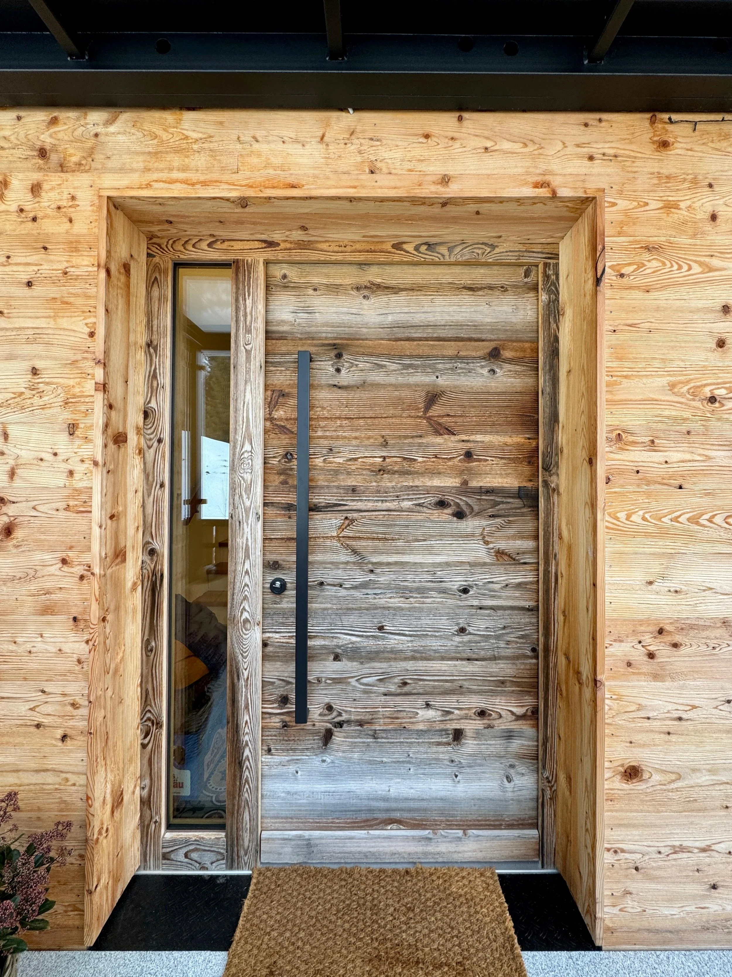 Moderner Eingang mit Holzrahmen und Glaselementen, schwarze Türklinke und Türgriff, begrüntes Beet links, brauner Fußmatte vor der Tür.