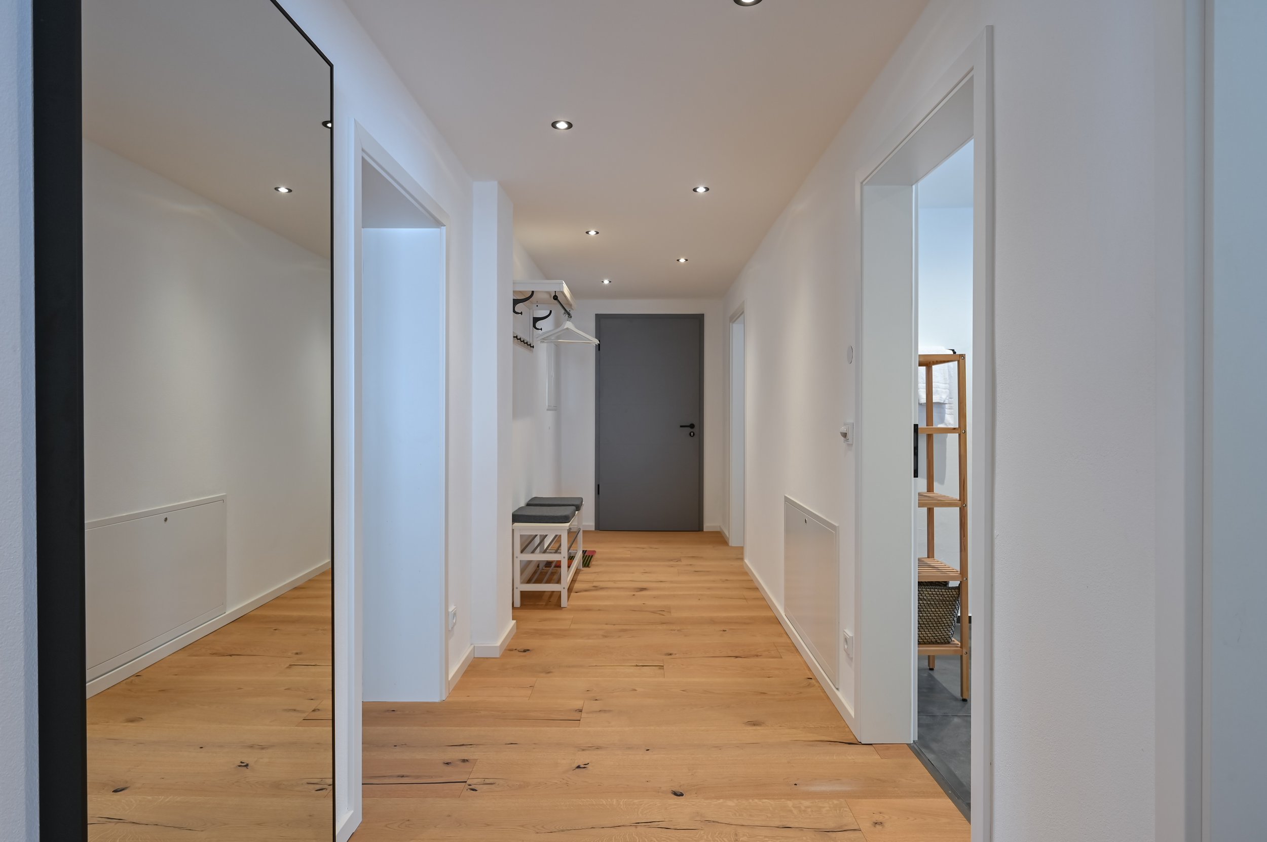 Ein heller Flur mit Holzfußboden, weißen Wänden und mehreren Türen, eine mit einem großen Spiegel