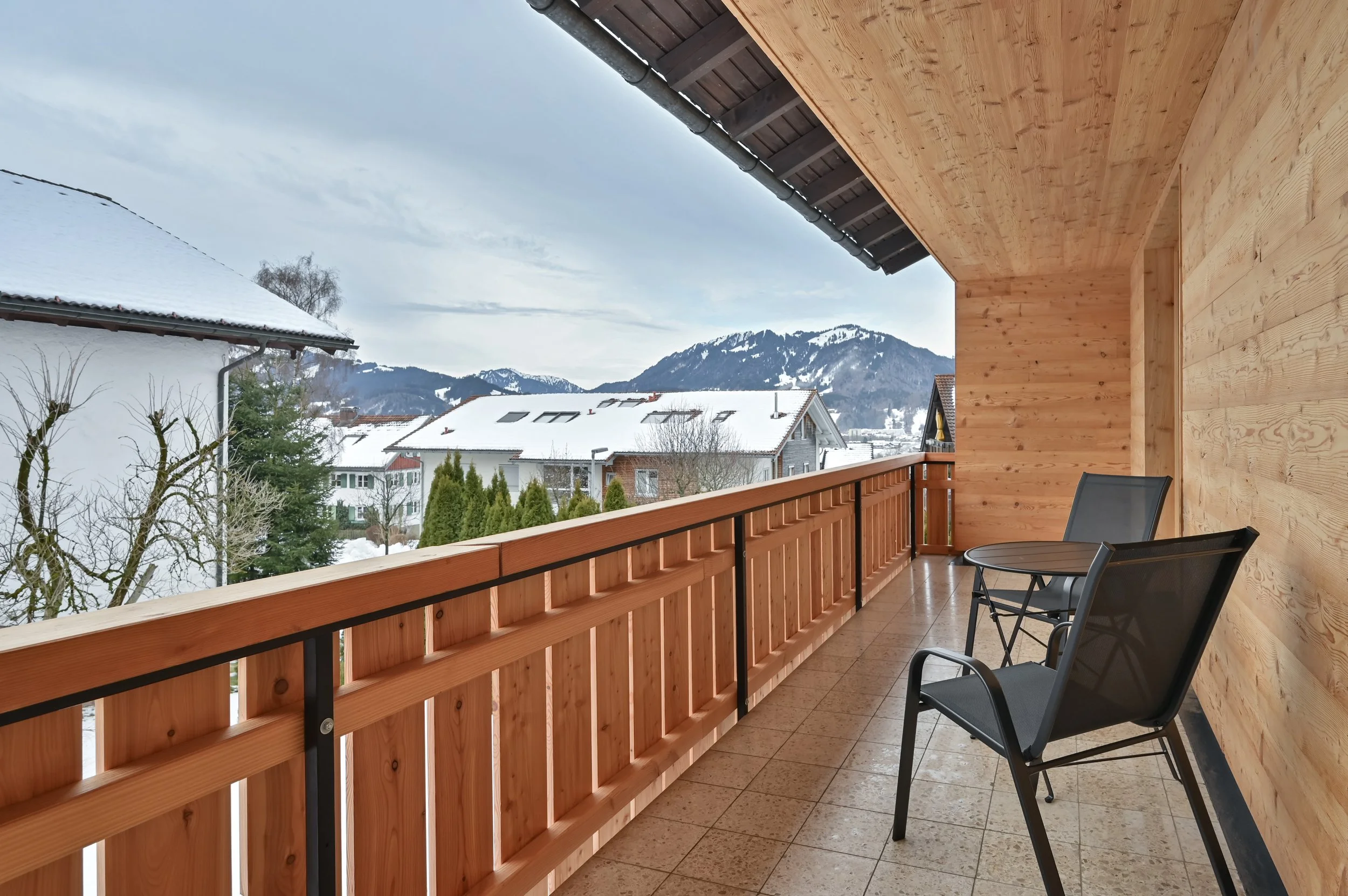 Blick von einem Balkon in einer winterlichen ländlichen Gegend mit verschneiten Dächern und Bergen im Hintergrund, Holzveranda mit zwei schwarzen Stühlen und einem kleinen Tisch.