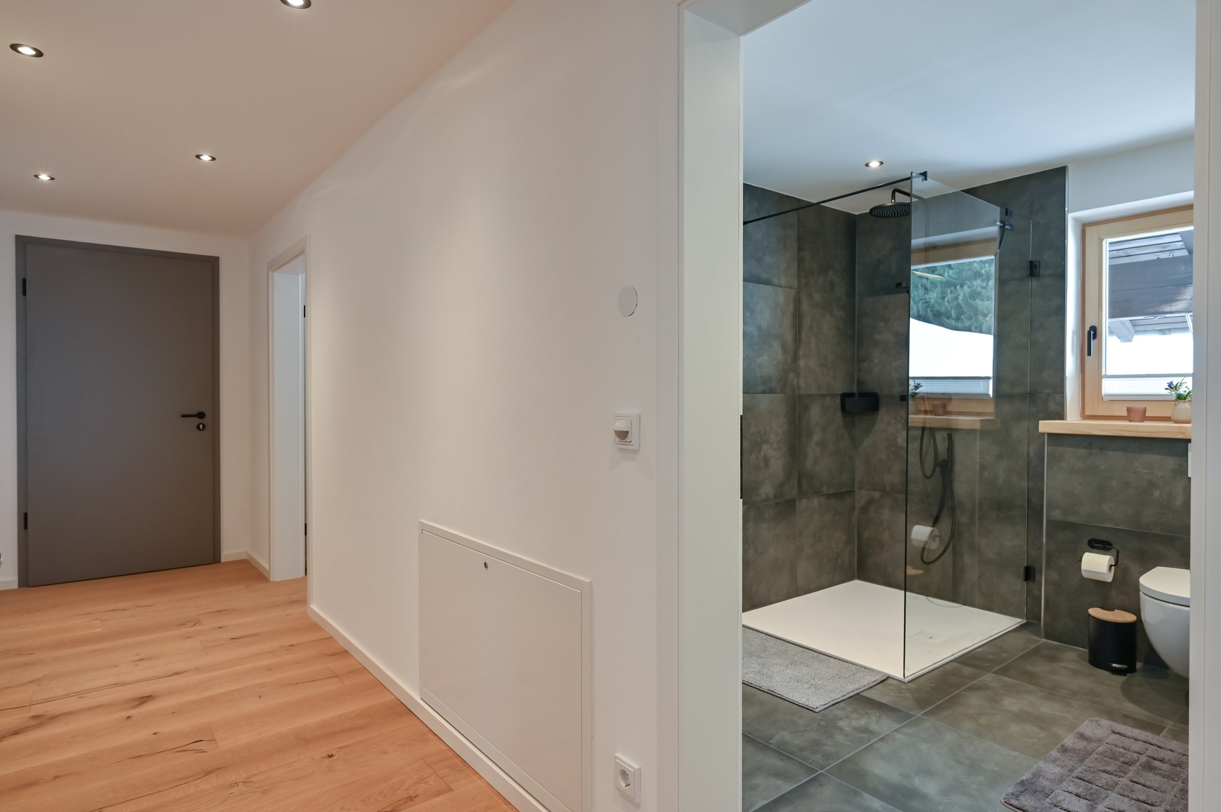 Innenaufnahme eines Flurs mit hellem Holzfußboden und weißen Wänden, links eine graue Tür, rechts eine offene Badezimmerdusche mit dunklen Fliesen, einer Dusche, WC und zwei Fenstern.