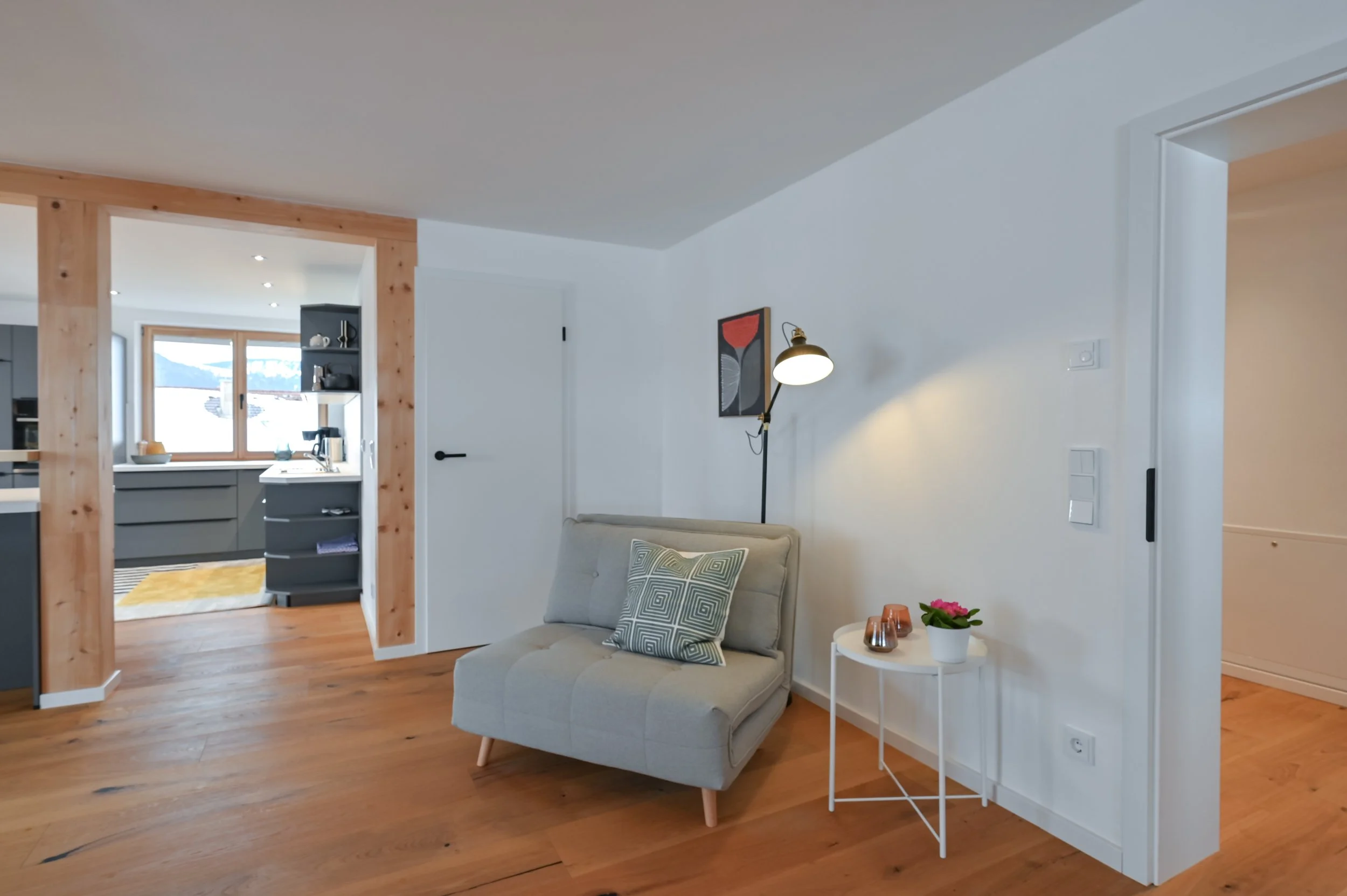 Innenraum eines Wohnzimmers mit Holzfußboden, einem grauen Sessel, einer weißen Beistelltischchen mit Topfpflanze und Kerzen, einer Stehlampe, weißen Wänden und einer Tür sowie einem Blick in die Küche im Hintergrund.