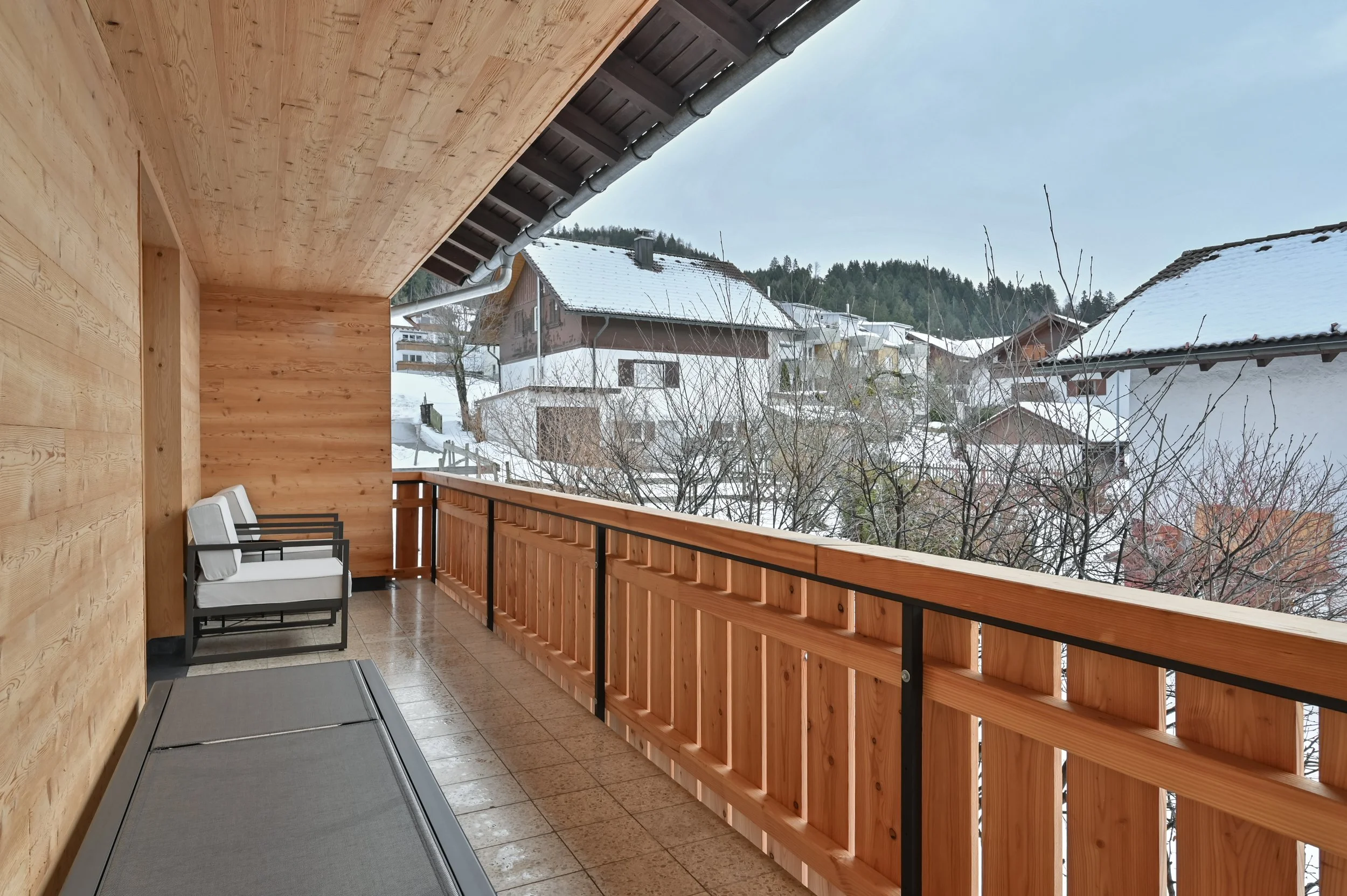 Balkon mit Holzverkleidung, zwei Stühle, Schnee bedecktes Haus und Bäume im Hintergrund.