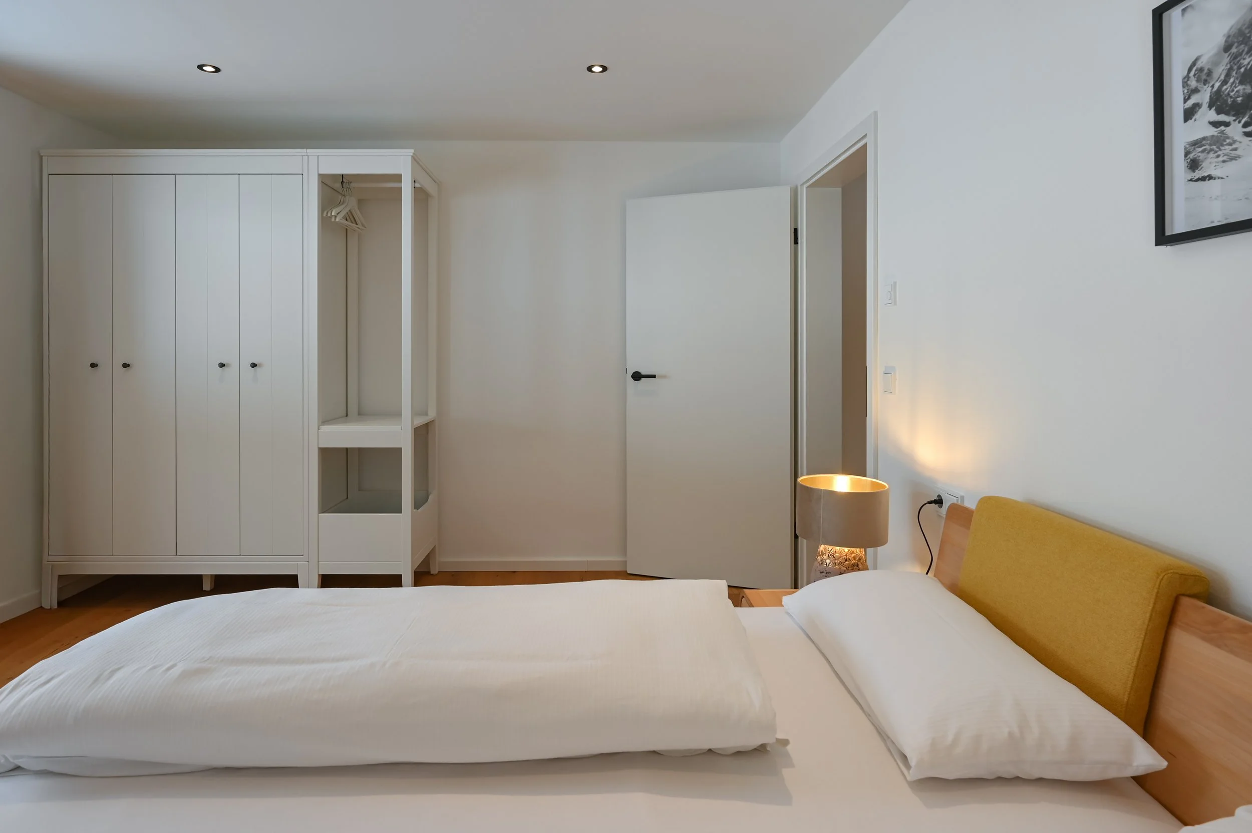 Ein modernes, minimalistisch eingerichtetes Schlafzimmer mit einem weißen Bett, gelbem Kopfteil, Nachttisch mit Stehlampe, weißem Kleiderschrank und offener Tür.