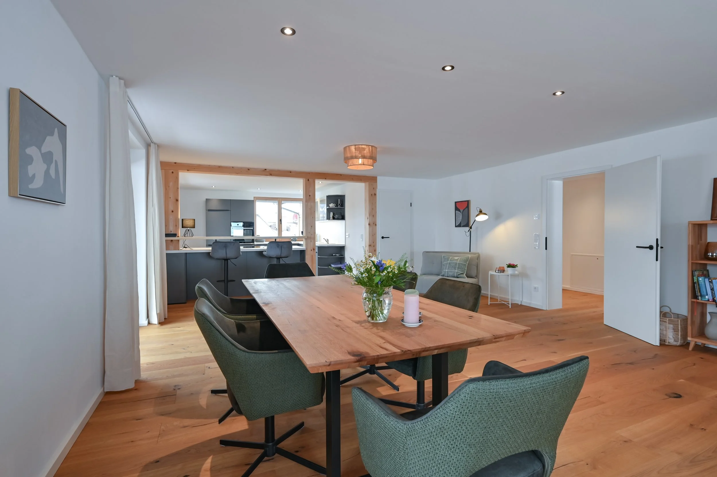 Offener Wohn- und Essbereich mit Holztisch, grünen und schwarzen Sesseln, moderner Küche im Hintergrund, Dekorationen, und Holzfußboden.