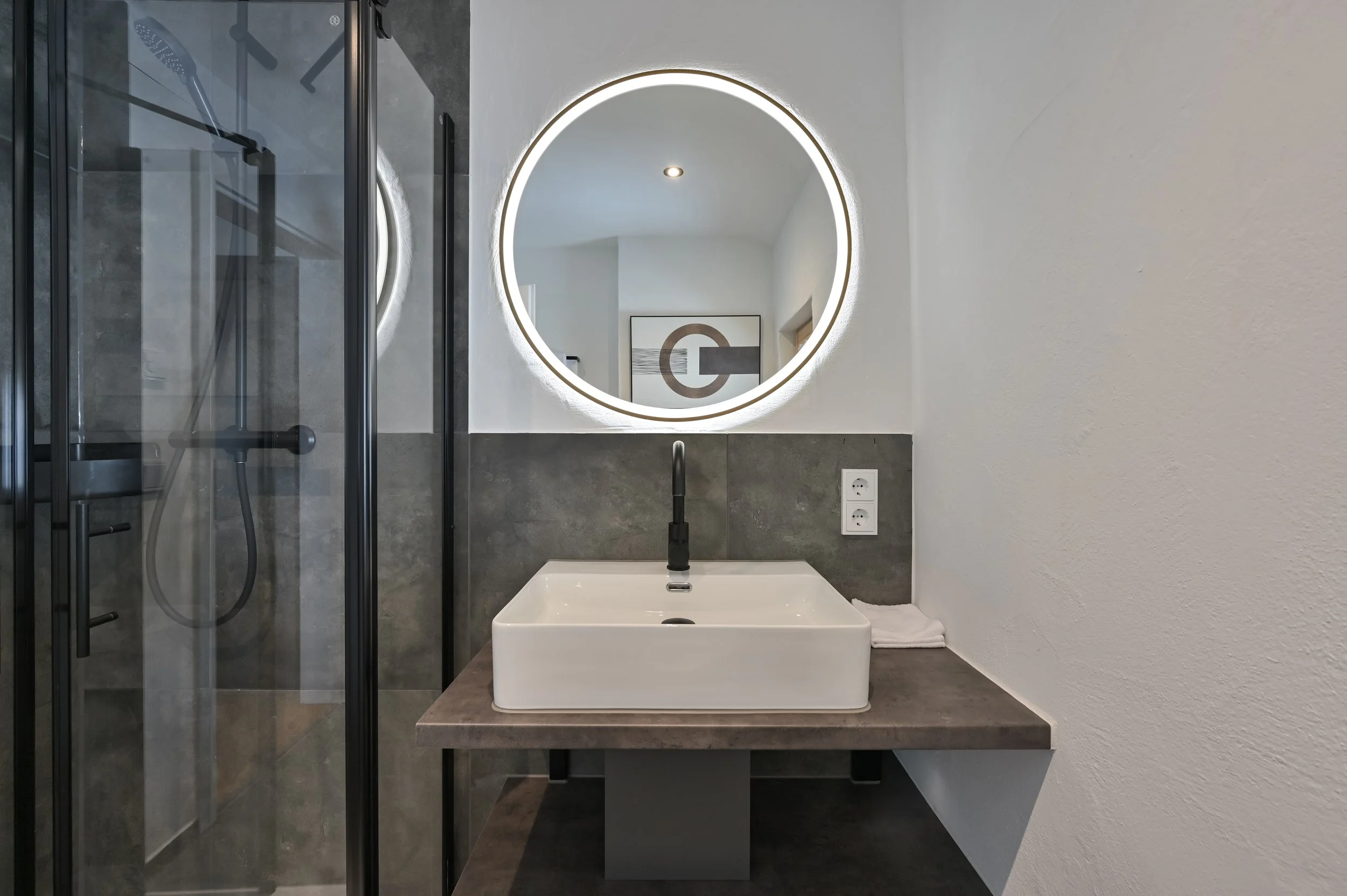 Moderner Badezimmerbereich mit Waschtisch, rundem beleuchteten Spiegel und separater Duschbereich mit schwarzer Glastür.