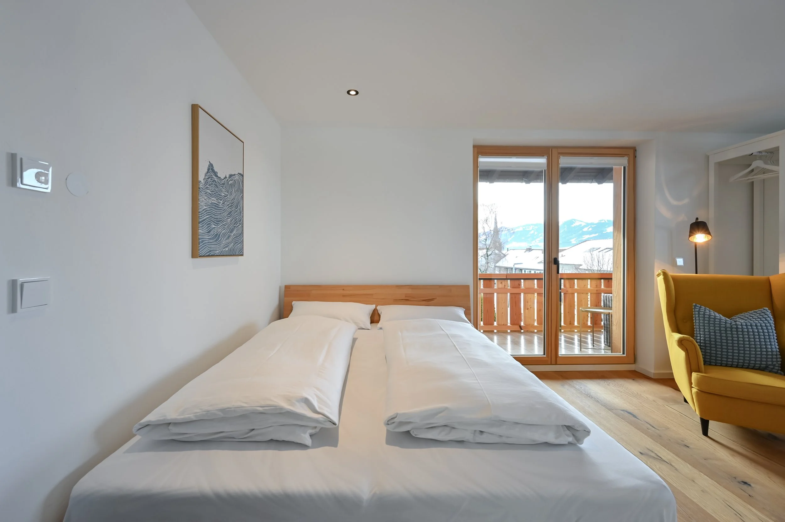Schlafzimmer mit Doppelbett, Holzbalkon mit Aussicht auf Berge, gelbe armlehnstuhl, Wandbild mit Wellenmuster, Parkettboden, Fenster mit Holzrahmen und Blick auf Winterlandschaft