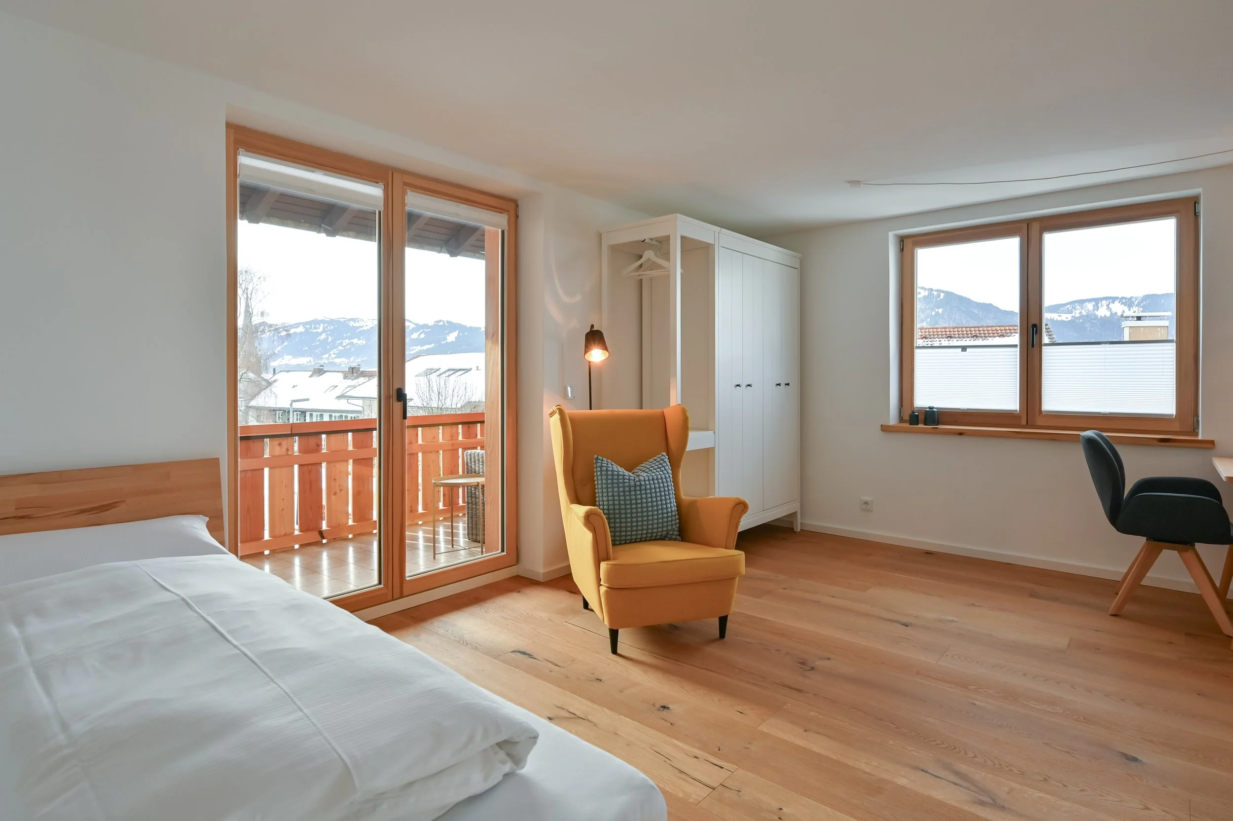 Ein modern eingerichtetes Apartment mit Holzboden, einem offenen Balkon, einem gelben Sessel, einem weißen Kleiderschrank, einem Schreibtisch und einem Fenster mit Blick auf Berge und Schnee.