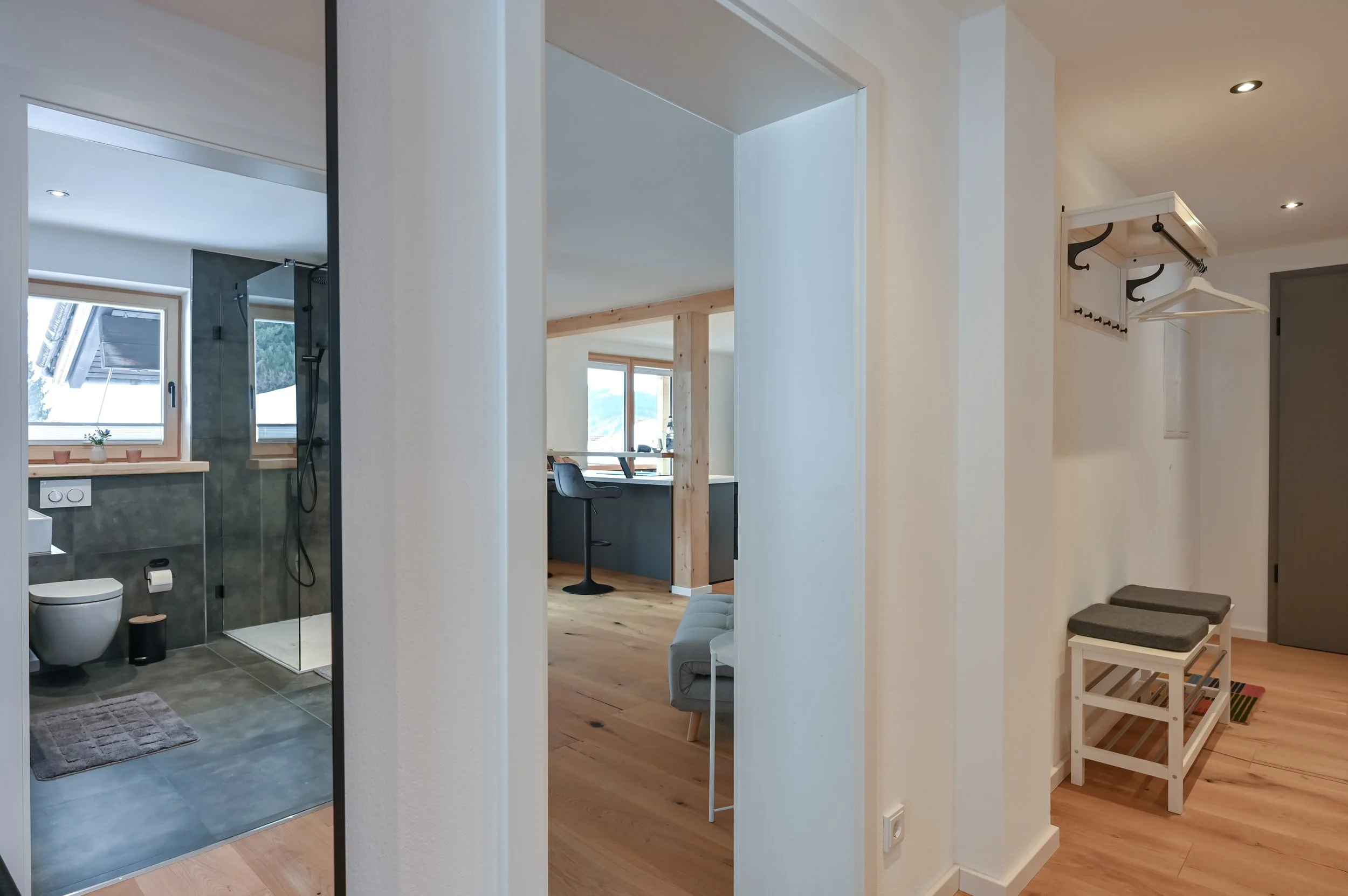 Innenansicht eines Hauses mit Blick auf Badezimmer, Küche und Flur. Das Badezimmer hat eine Toilette und eine Dusche. Die Küche ist modern mit Barhockern. Der Flur hat eine Sitzbank, Haken und eine Ablage mit Kleiderbügeln.