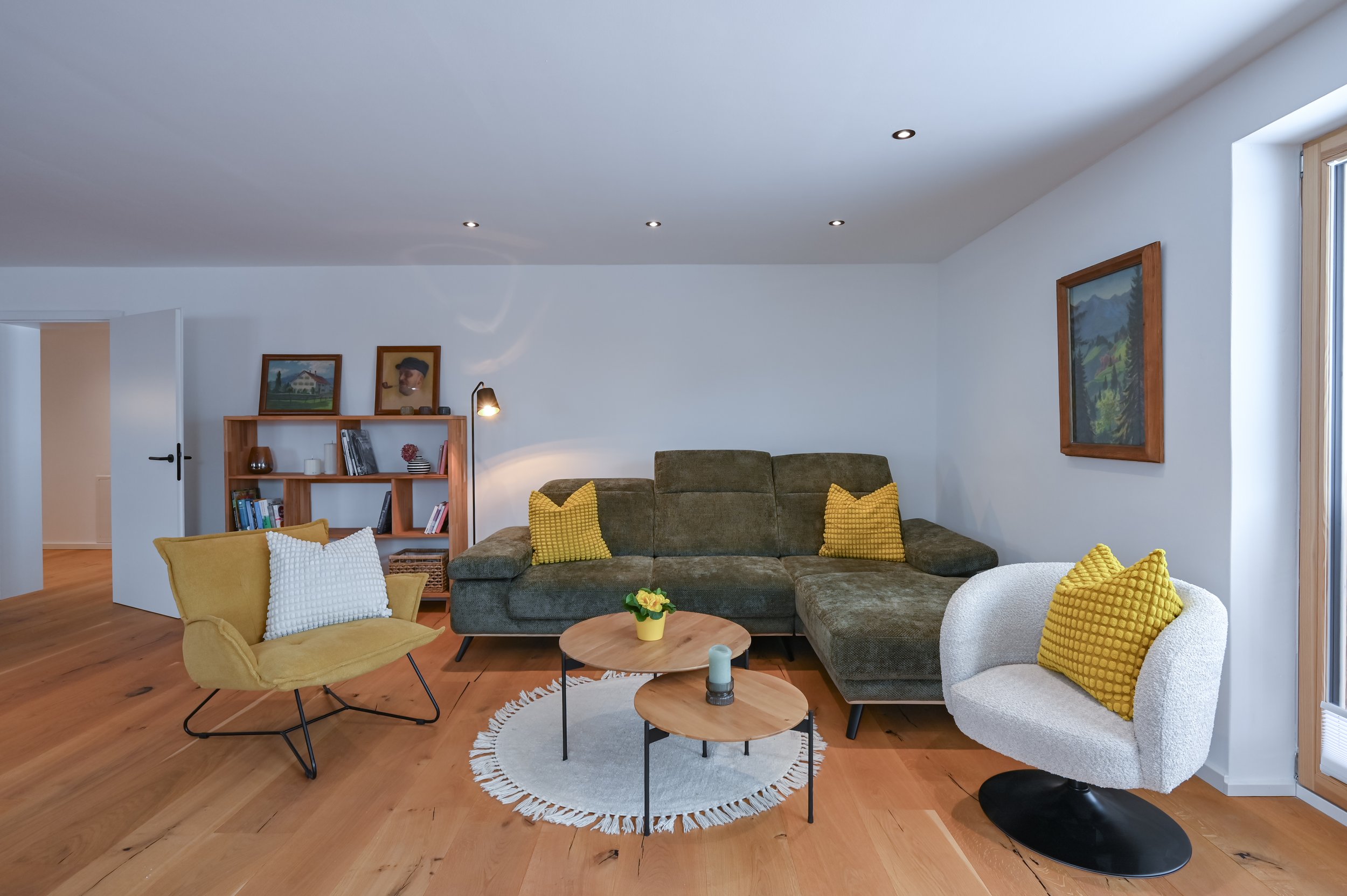 Wohzimmer mit grüner Couch, zwei Sessel mit gelben Kissen, Holztisch mit Dekoration, Bücherregal, Wandbild, Holzfußboden, weiße Wände, natürliches Licht von Fenster und Deckenleuchte.