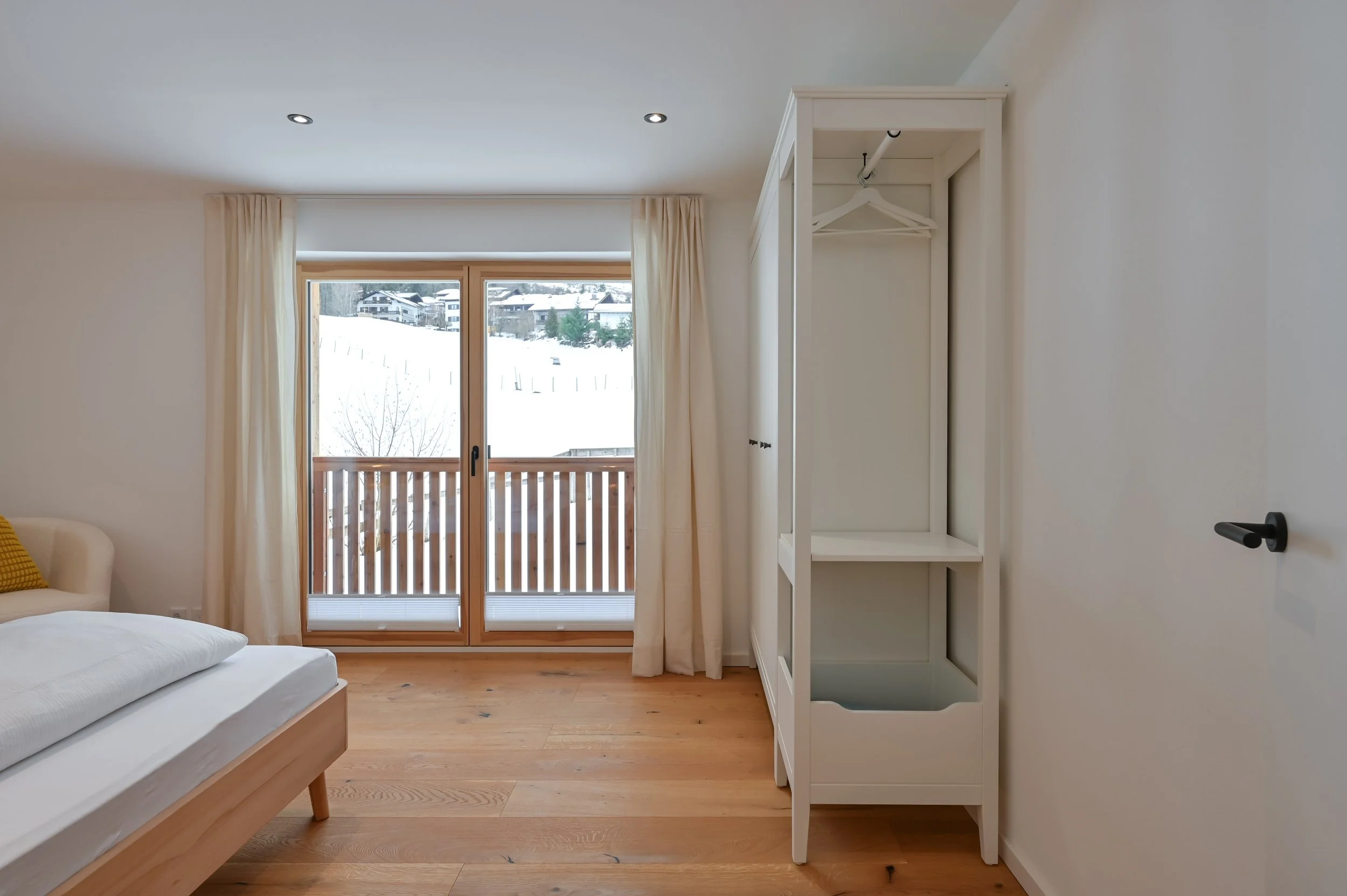 Ein Schlafzimmer mit Holzfußboden, einem weißen Bett, einem kleinen Sofa mit gelbem Kissen, einem Balkon mit Holzgeländer und einem Schneelandschaft im Hintergrund, sowie einem weißen Kleiderschrank an der Wand.