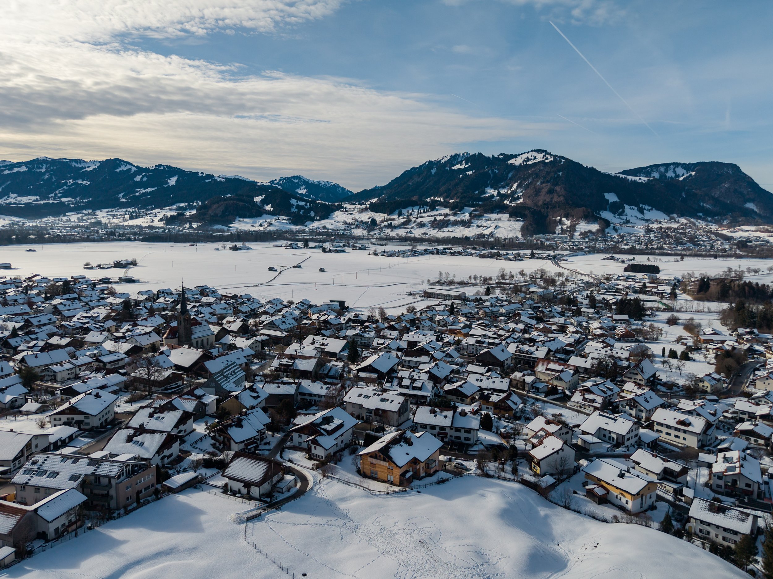 Eine verschneite Stadt im Tal mit vielen Häusern und einer Kirche im Zentrum, umgeben von schneebedeckten Feldern und Bergen im Hintergrund, bei klarem Himmel.
