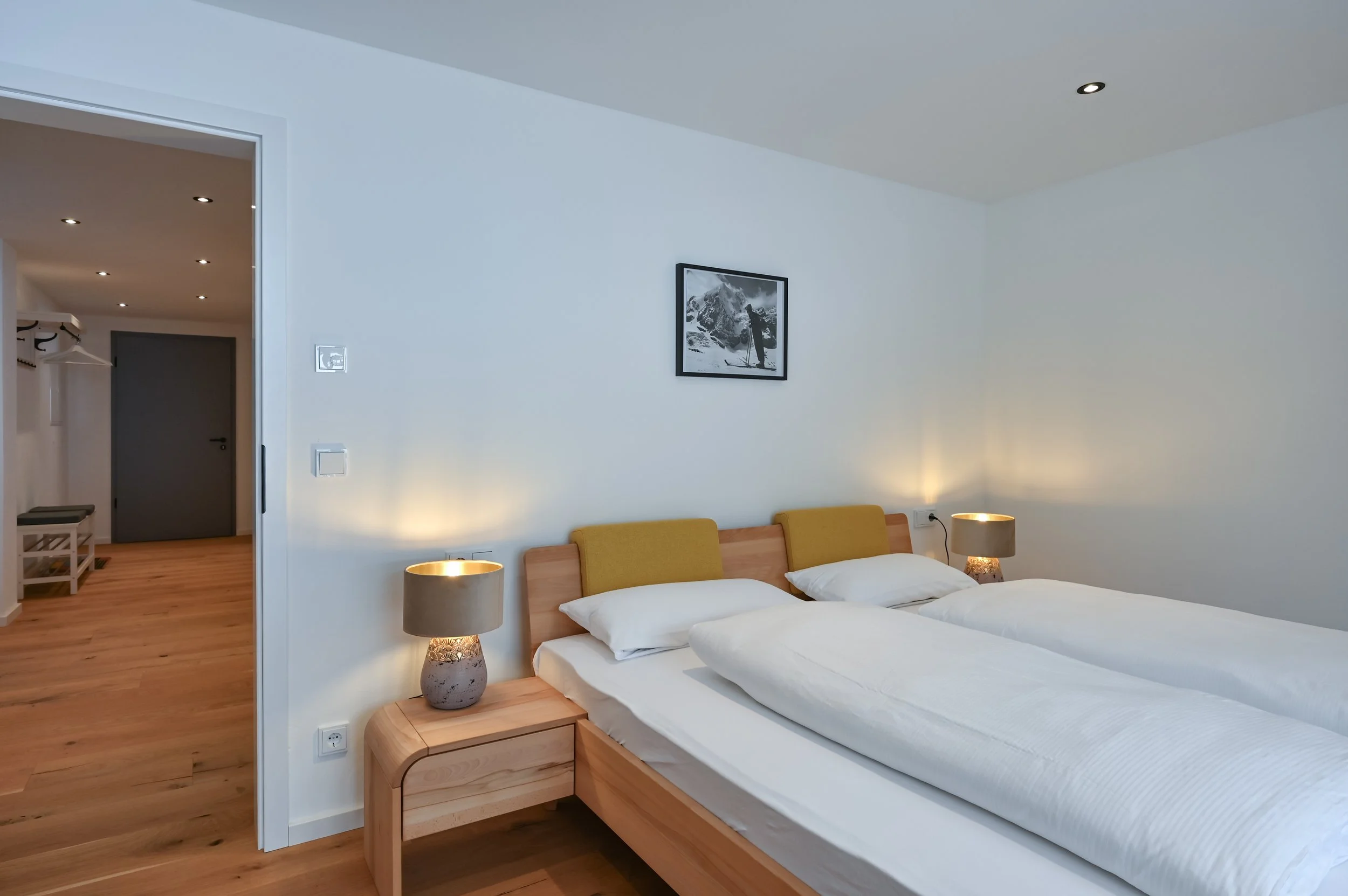 Schlafzimmer mit Doppelbett, Bettwäsche, Nachttischen mit Lampen, Wandbild, Holzboden, benachbarter Flur mit weißen Wänden, Garderobe und Tür.
