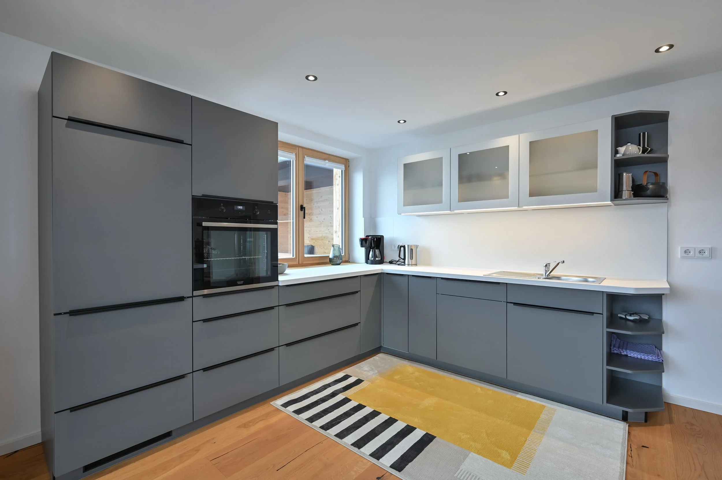 Moderne Küche mit grauen Schränken, weißen Arbeitsflächen, einem Herd, einem Kaffeeautomaten, einem Wasserkocher und einem Fenster mit Blick nach draußen.