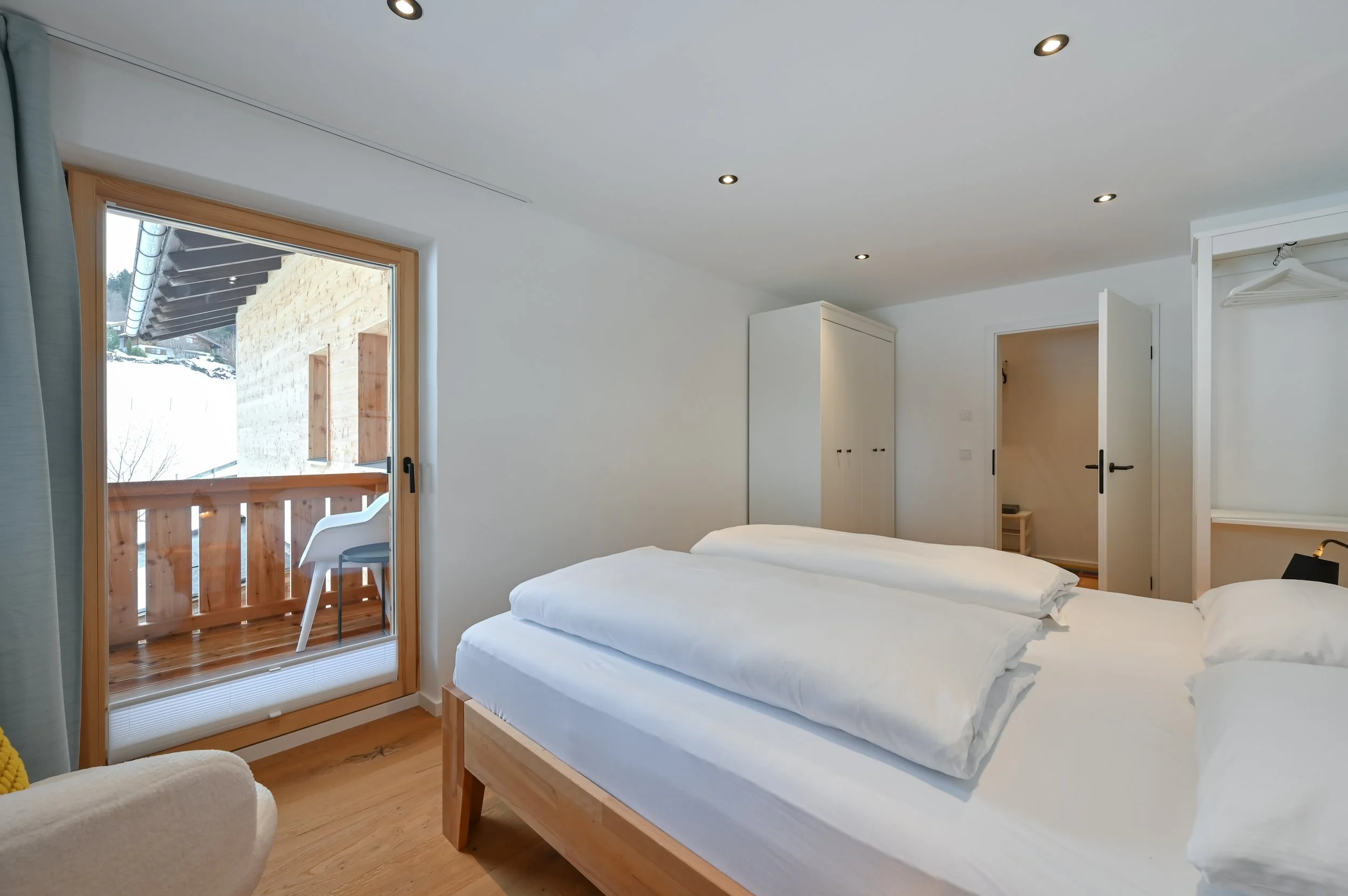 Schlafzimmer mit weißem Bett, Holzfußboden, Balkon mit Holzgeländer und Plastikstuhl, offene Garderobe, weiße Wände, Deckenleuchten.