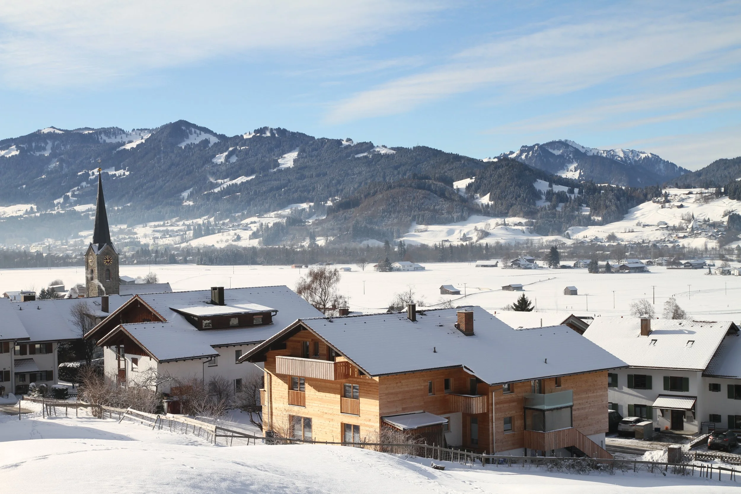 Häuser in einer verschneiten Landschaft mit Bergen im Hintergrund, eine Kirche mit hohem Turm im Vordergrund.