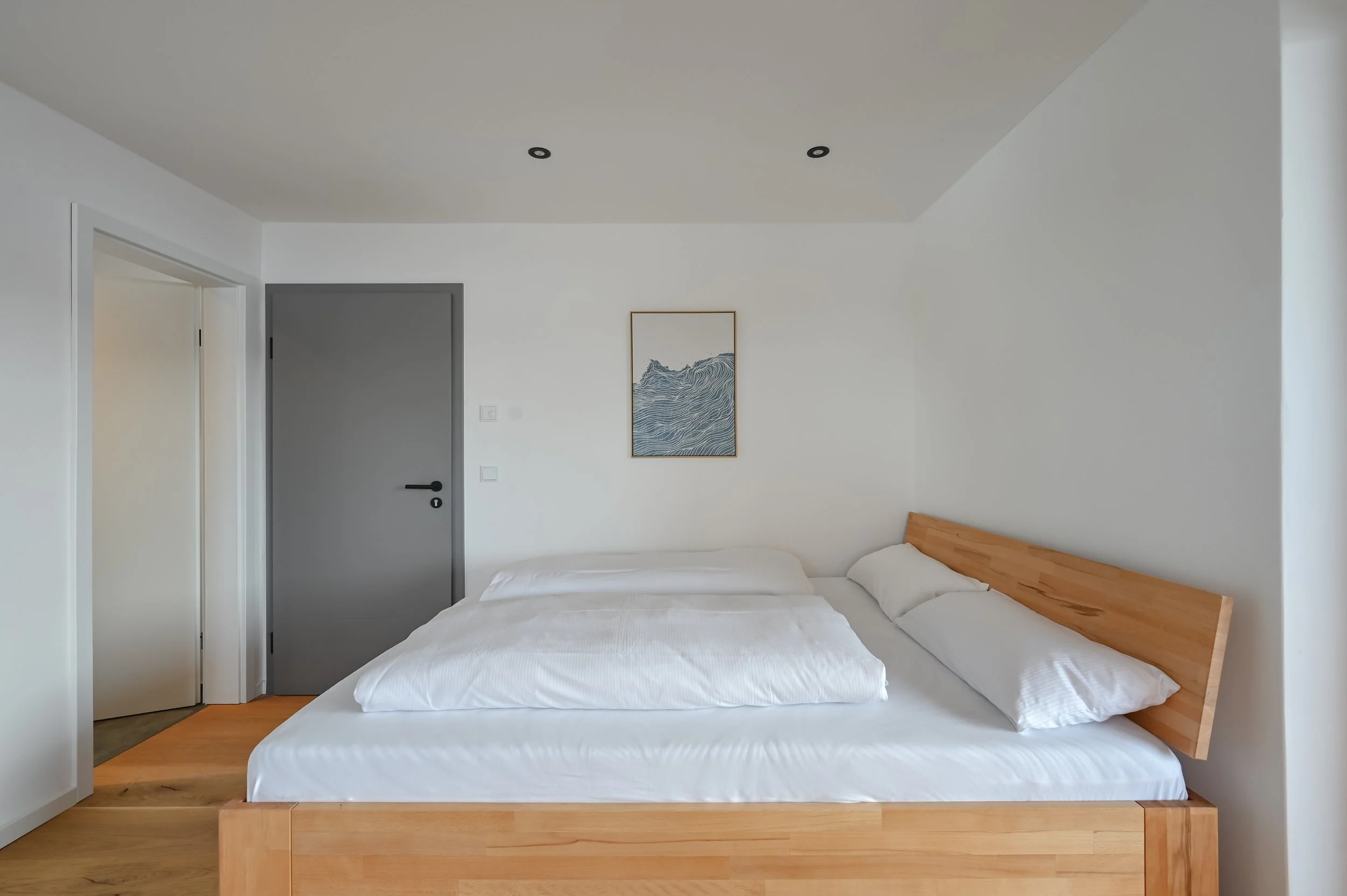 Modernes Schlafzimmer mit hellem Holzbett, weißen Bettwäsche, Wandbild über dem Bett, graue Tür, weiße Wände, Deckenleuchte.