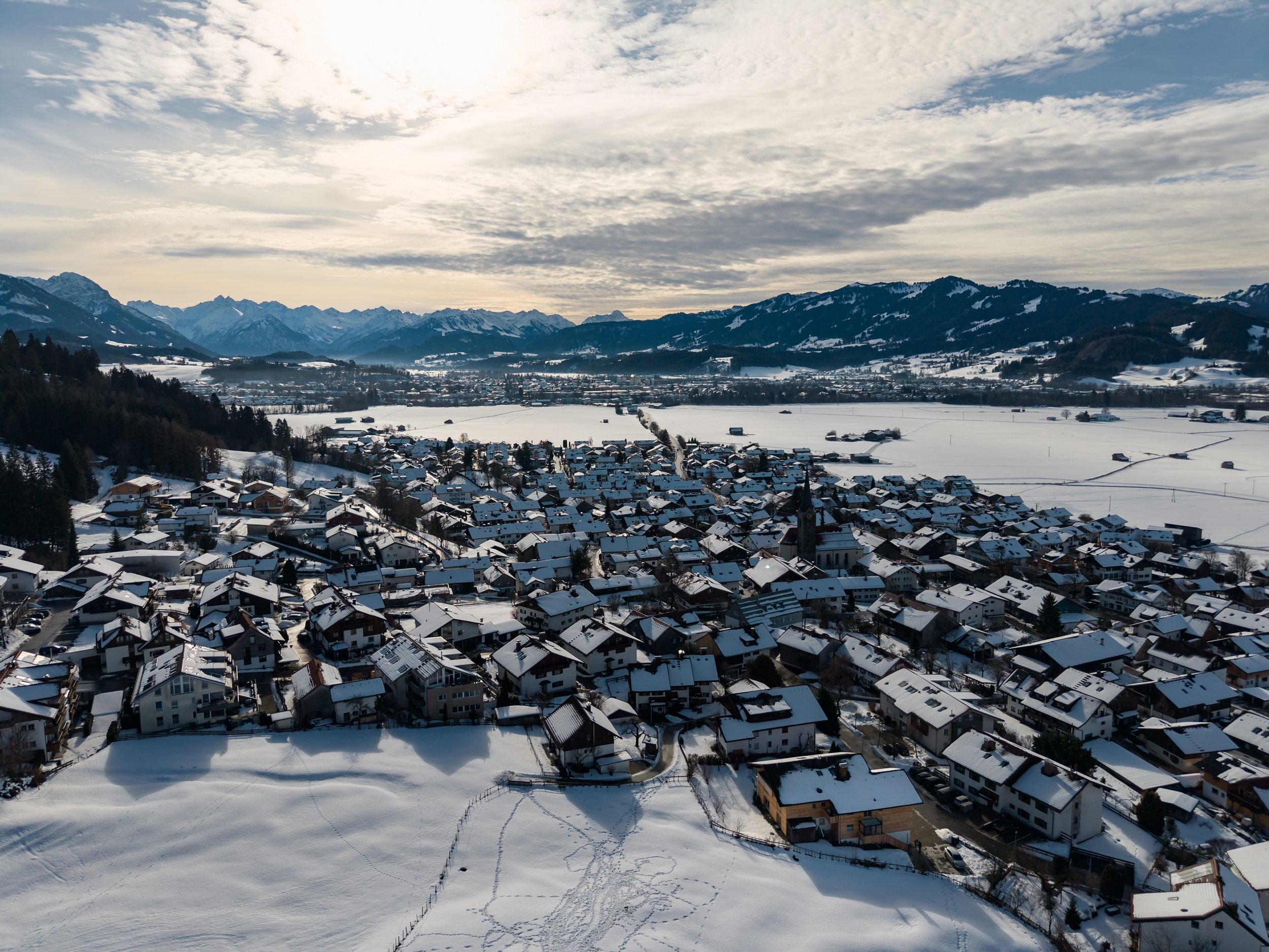 Luftaufnahme eines verschneiten Dorfes in einer bergigen Region bei bewölktem Himmel.