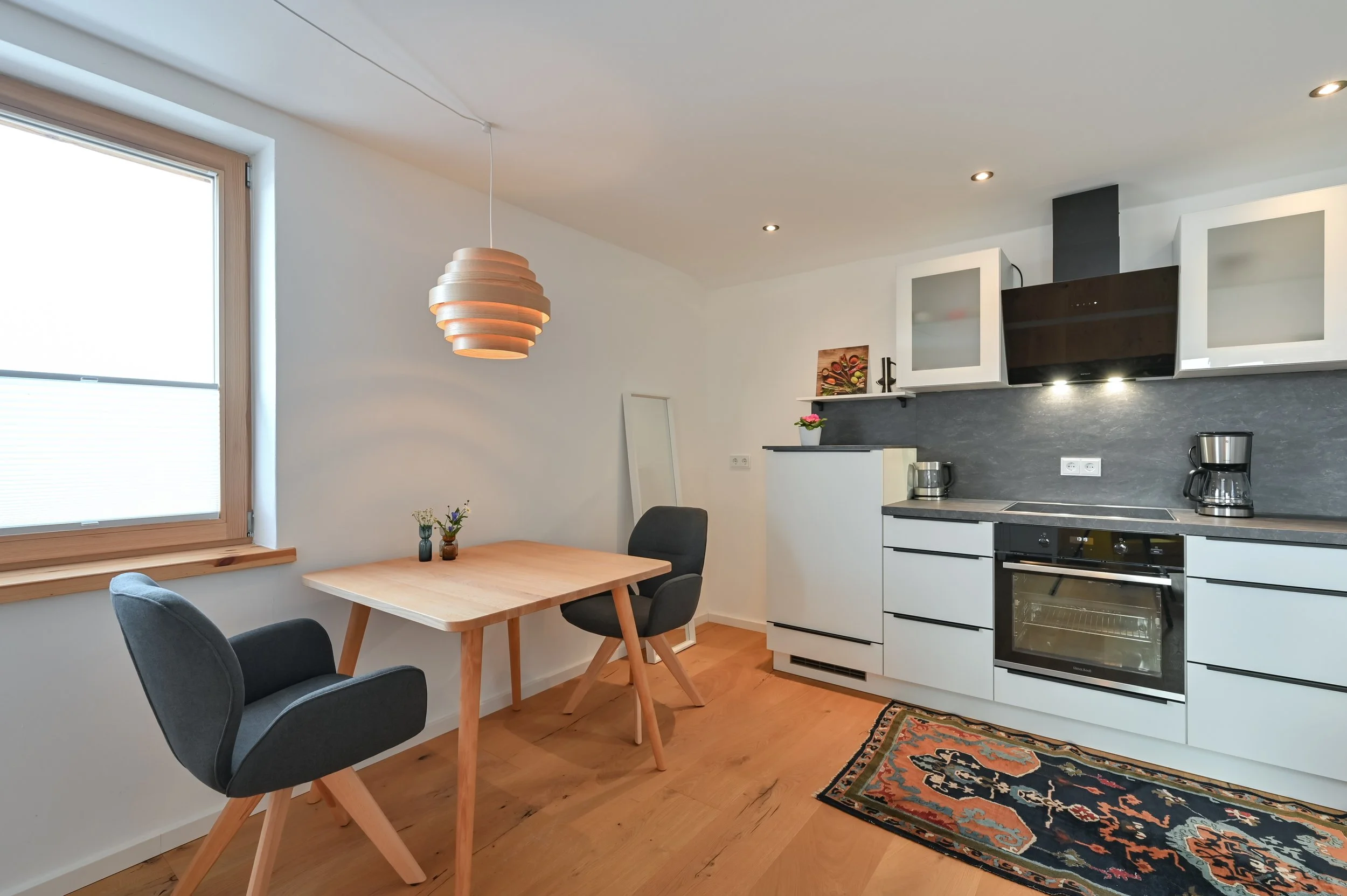 Moderne Küche und Essbereich mit einem kleinen Holztisch, zwei dunklen Stühlen, weißen Küchen Schränken, dunklem Backofen, Wasserkocher, Kaffeemaschine, Wanddekoration und einem Teppich auf einem Holzfußboden.