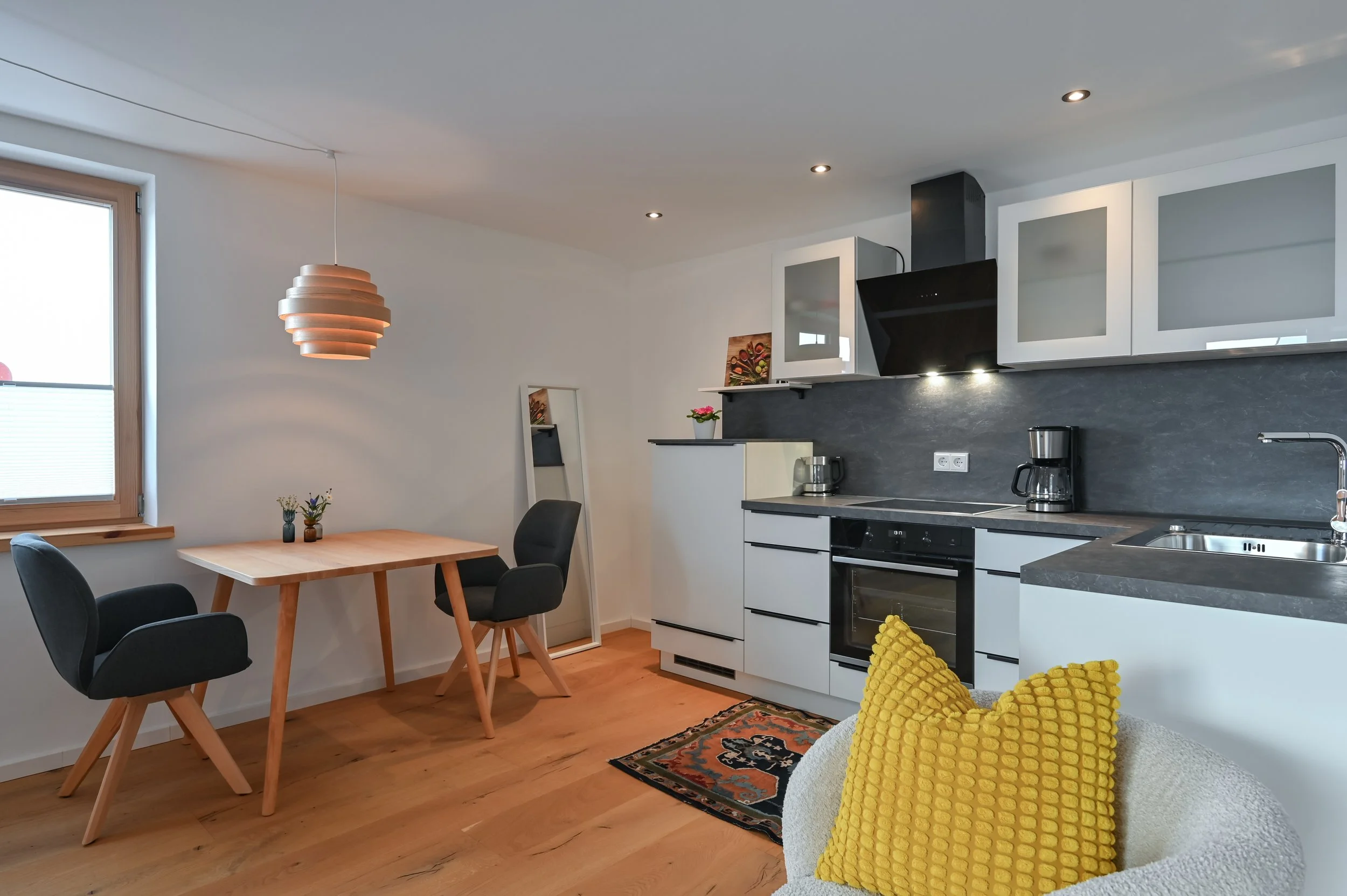 Offene Küche mit weißen Schränken, schwarzen Arbeitsplatten, Herd, Mikrowelle, Kaffeemaschine, Spüle und Andenken auf der Fensterbank, Esstisch mit drei schwarzen Stühlen, Holzparkett, Fenster mit Rollladen, Wandspiegel, Teppich mit Muster, mit gelbe