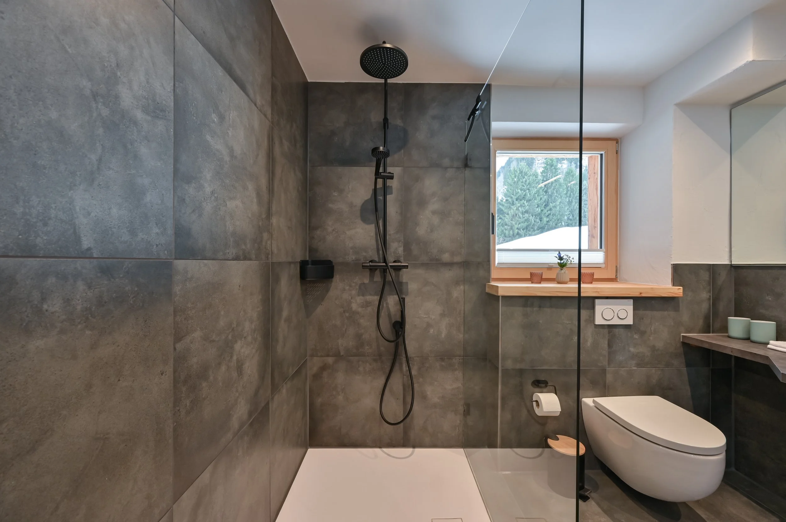 Modernes Badezimmer mit grauen Fliesen, begehbarer Dusche, Fenster mit Blick auf Schneelandschaft, Wand-WC, Holzregal und Spiegel.