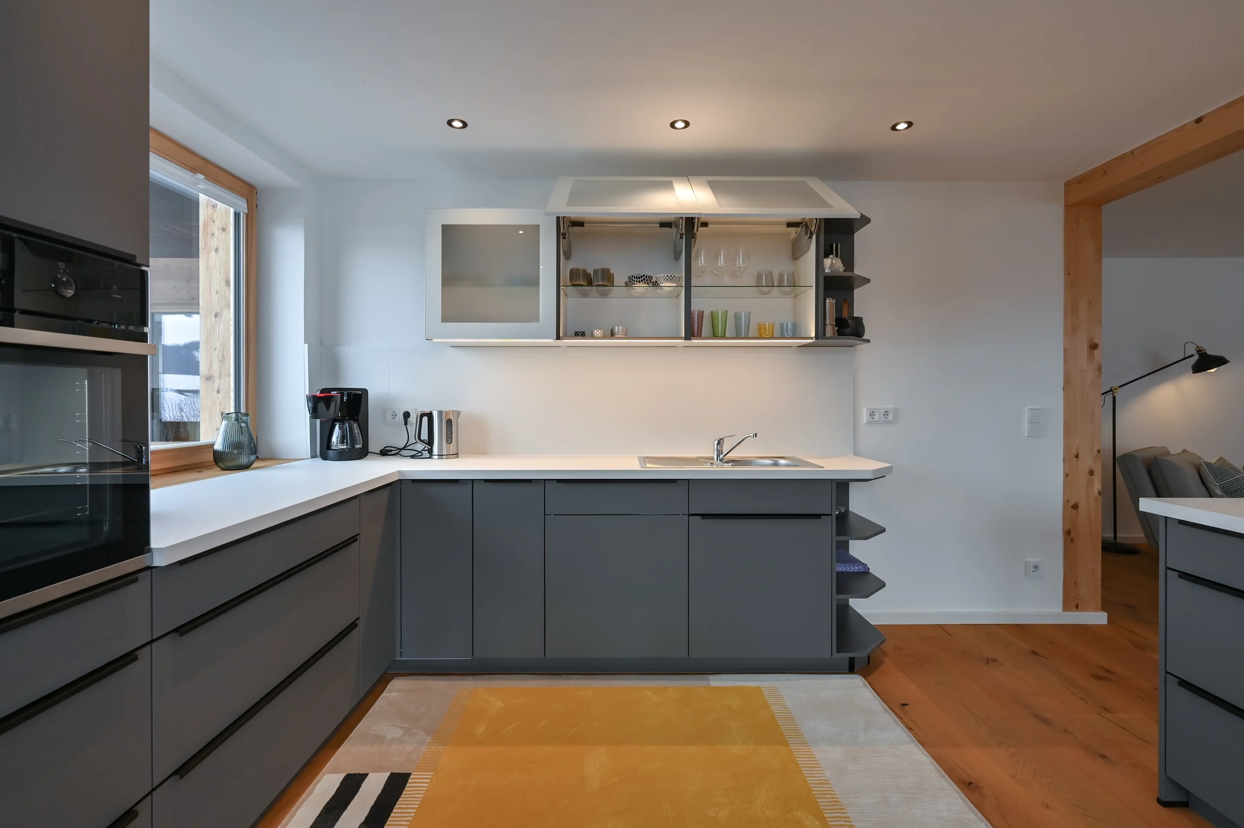 Moderne Küche mit grauen Schränken, weißen Arbeitsflächen, offenen Regalen mit Geschirr und Gläsern, Kaffeemaschine, Wasserkocher und Mikrowelle, neben einem Fenster mit Blick nach außen, Holzfußboden und türkiser Teppich.