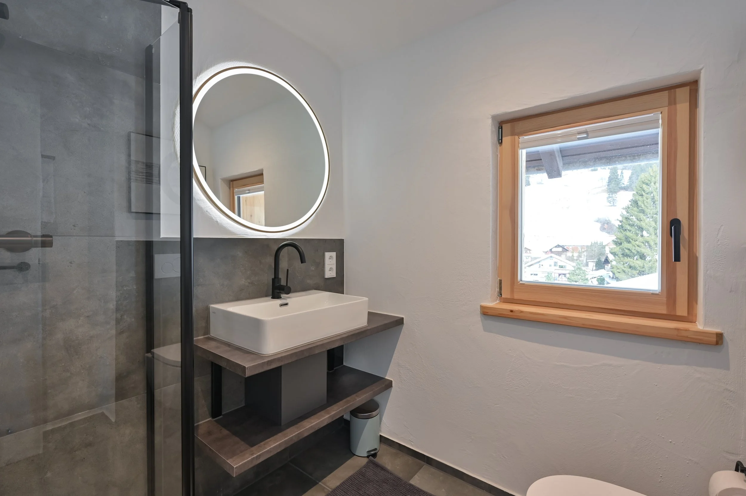 Modernes Badezimmer mit weißem Waschbecken, rundem beleuchtetem Spiegel, Holzfenster und grauer Fliese