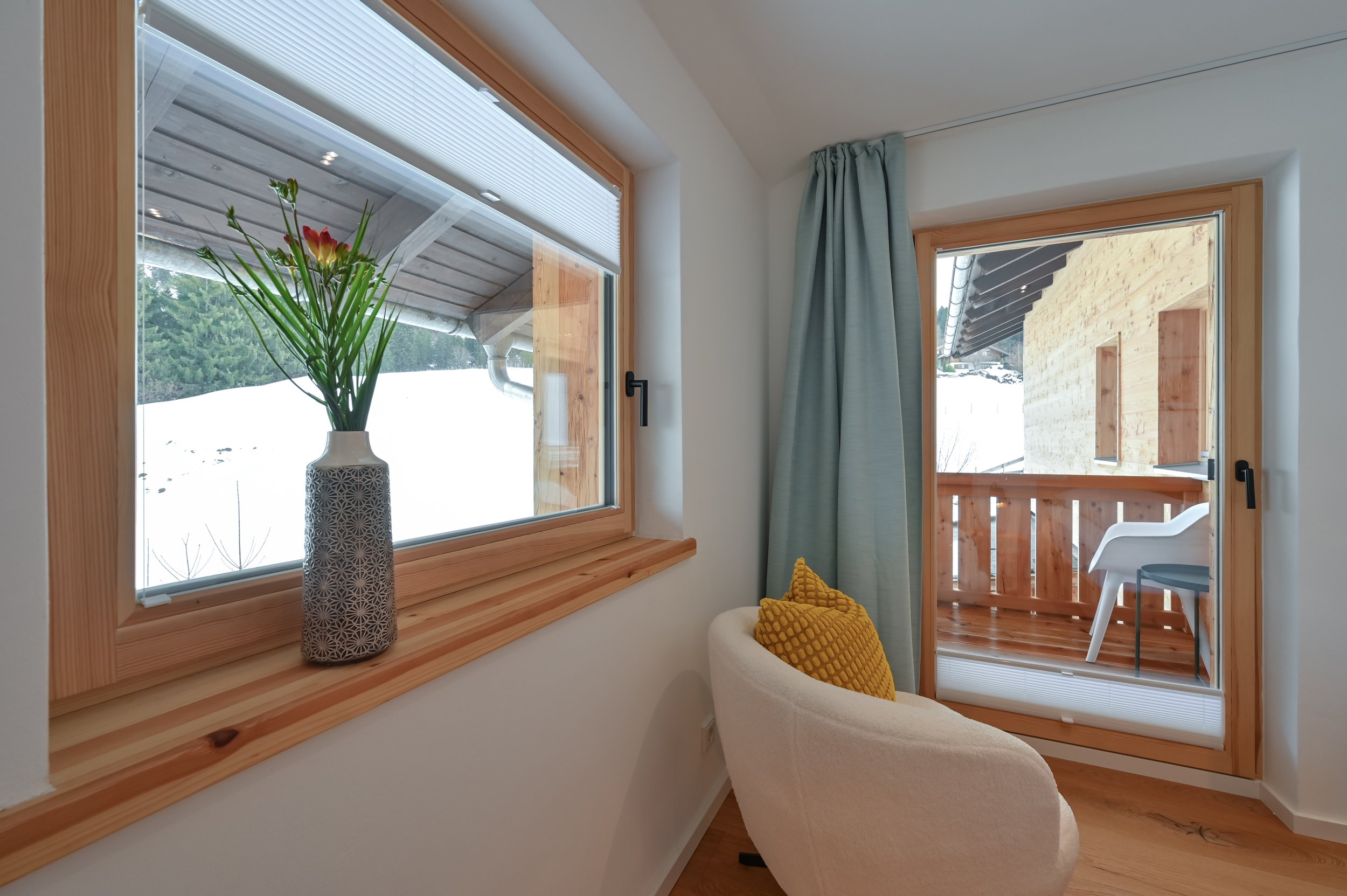 Innenraum eines Zimmers mit Fenster, Tür auf einen Balkon, eine weiße Sessel mit gelben Kissen, eine Vase mit Blumen auf der Fensterbank.