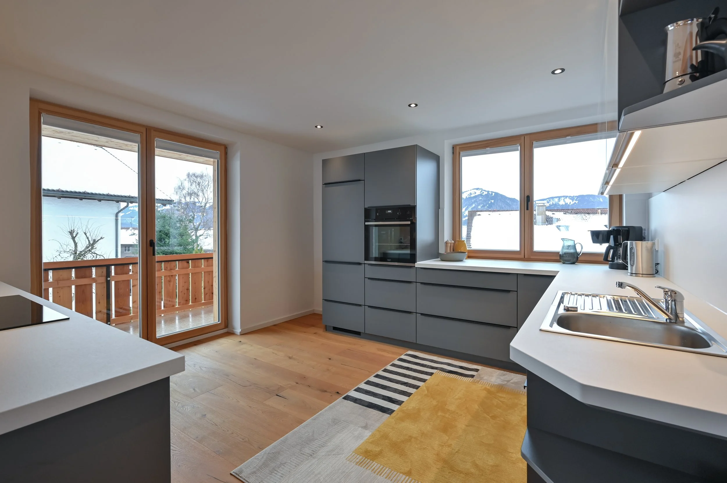Moderne Küche mit grauen Schränken, Holzfenstern, einem Herd, einer Spüle, Kaffeemaschine und Blick auf Berge und Schnee im Hintergrund.
