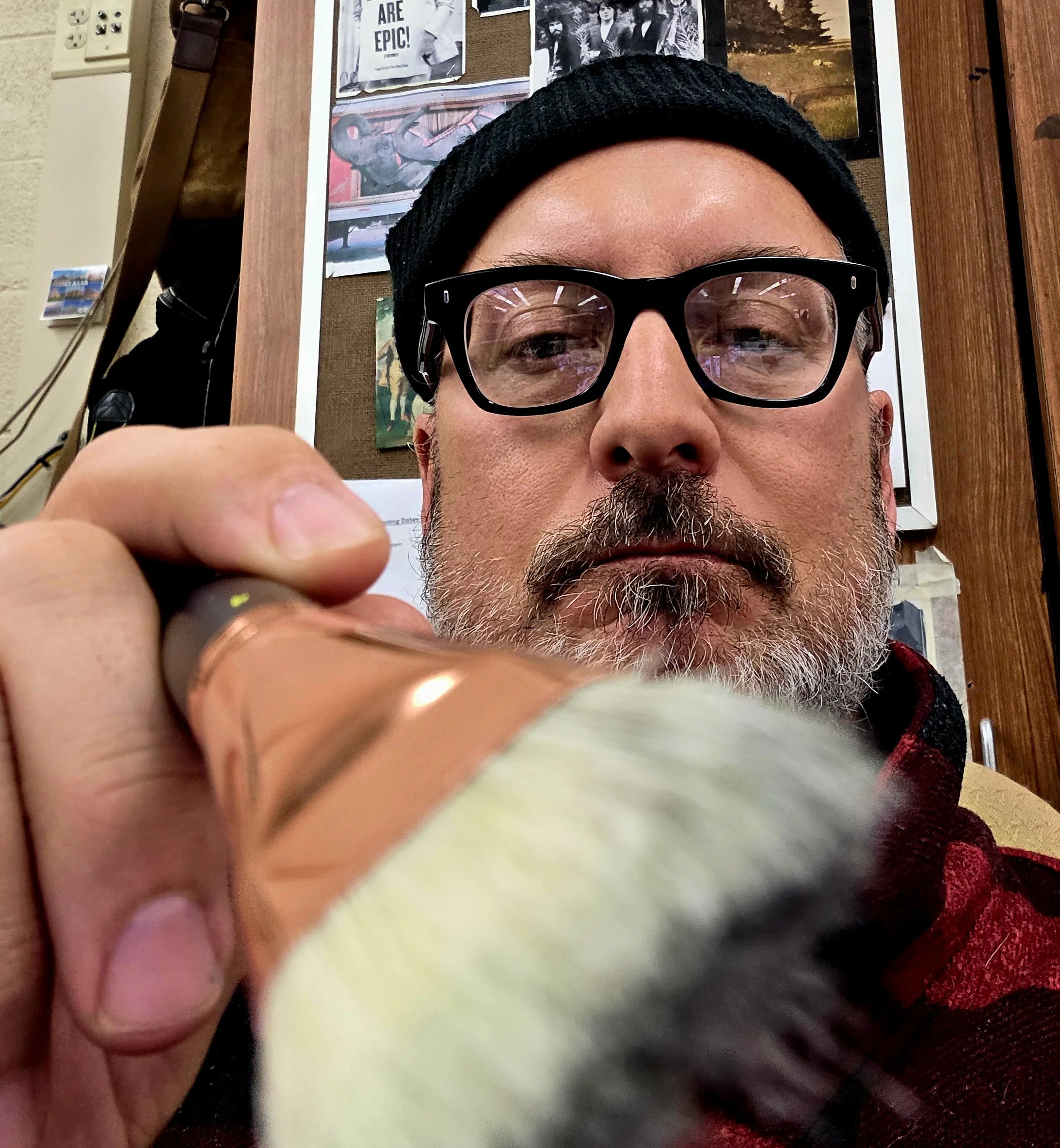 A man wearing black glasses, a black beanie, and a red shirt holds a paintbrush close to the camera, with a background of a bulletin board with photos and art.