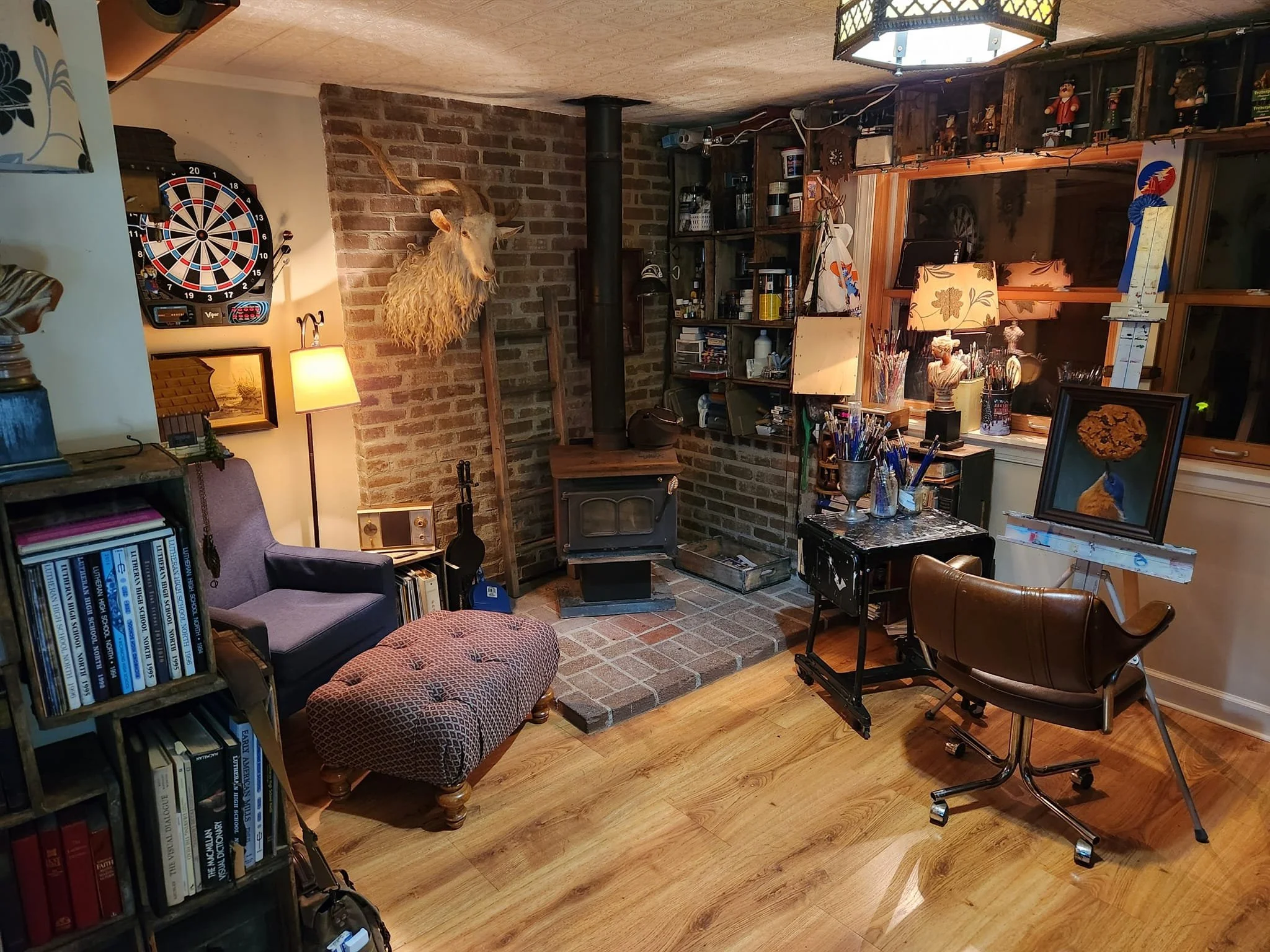 A cozy room with a brick fireplace, a mounted deer head, a purple armchair, and an ottoman, with a dartboard on the wall and various knick-knacks and books on shelves and tables, illuminated by warm lighting.