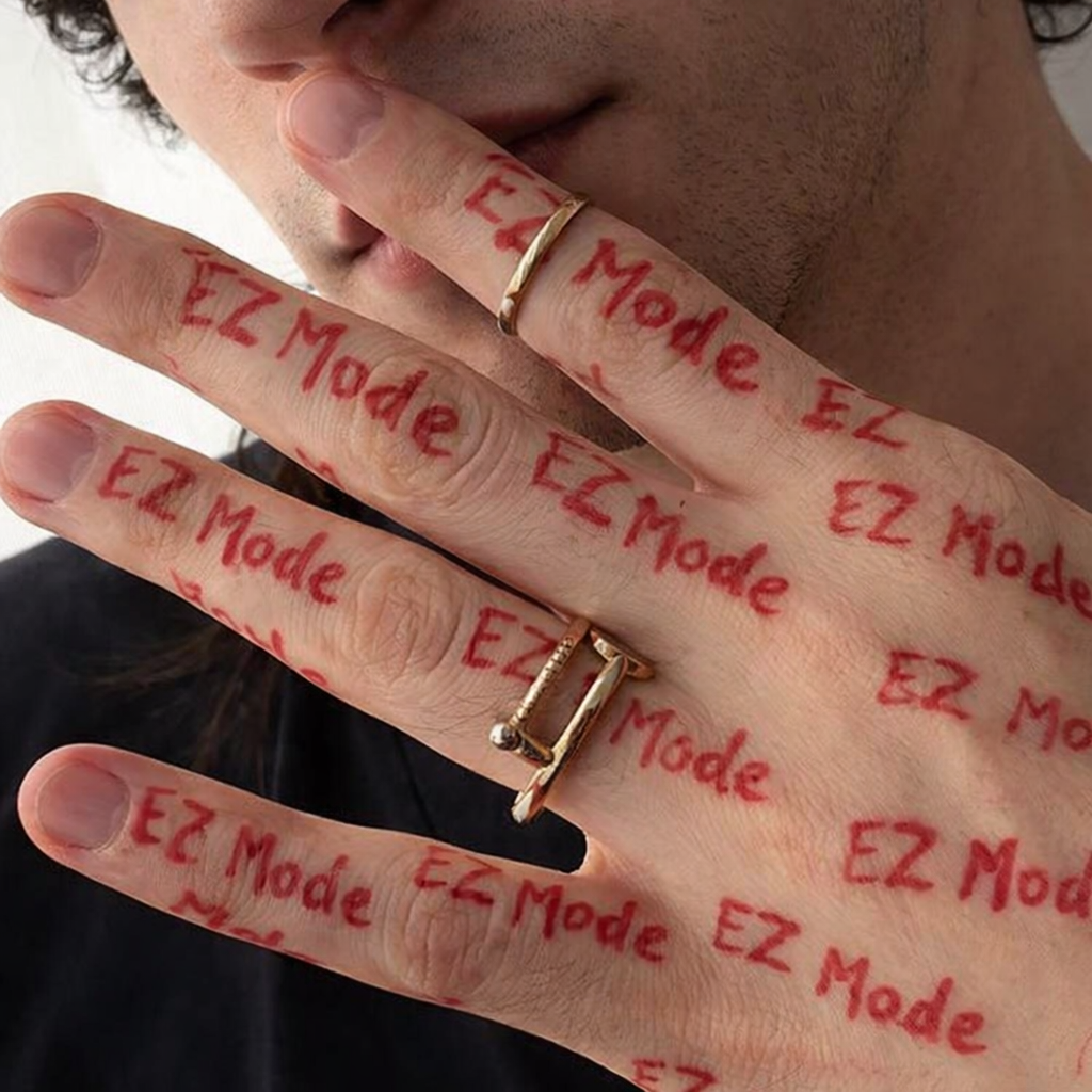 Person holding up his hand with fingers displaying the words 'EZ Mode' written repeatedly in red marker. The person is wearing rings and has a slight smile.
