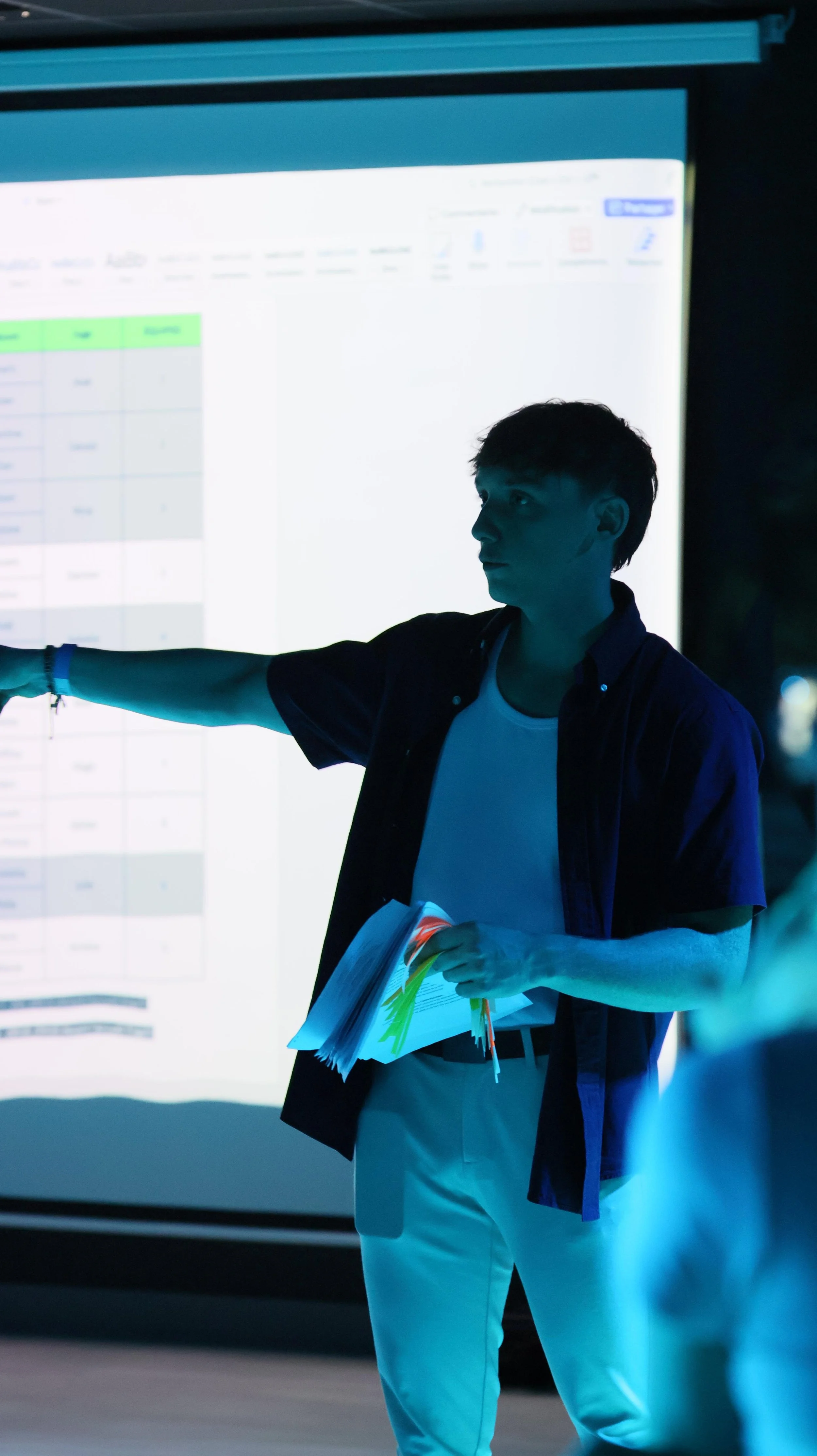 Un jeune homme en train de donner une présentation devant un tableau blanc ou un écran lumineux dans une salle sombre.