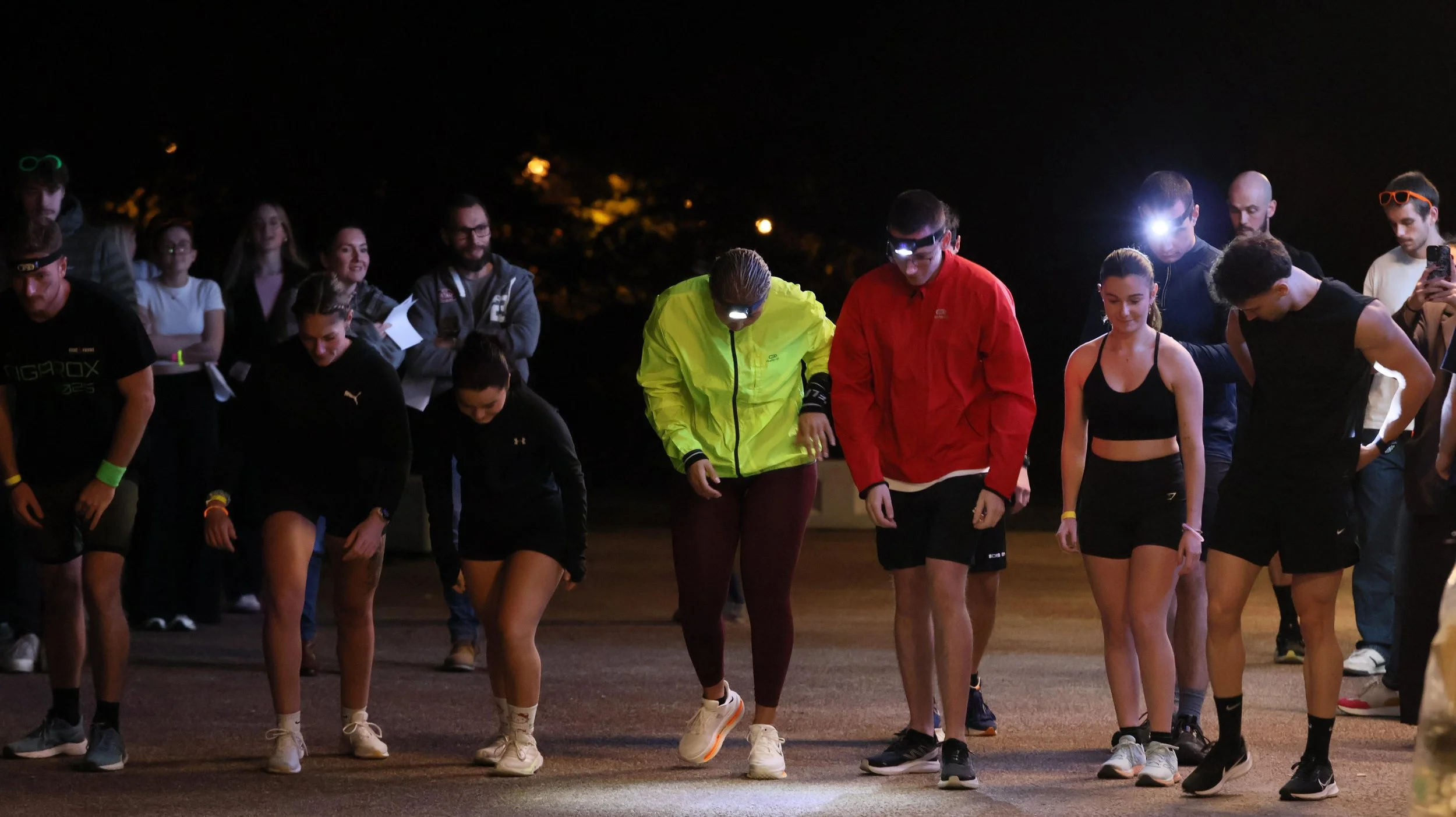 groupe de jeunes participants à une course nocturne, portant des vêtements de sport, certains avec des lampes frontales, en train de marcher ou de commencer une course sur une route. Hyrox nocturne