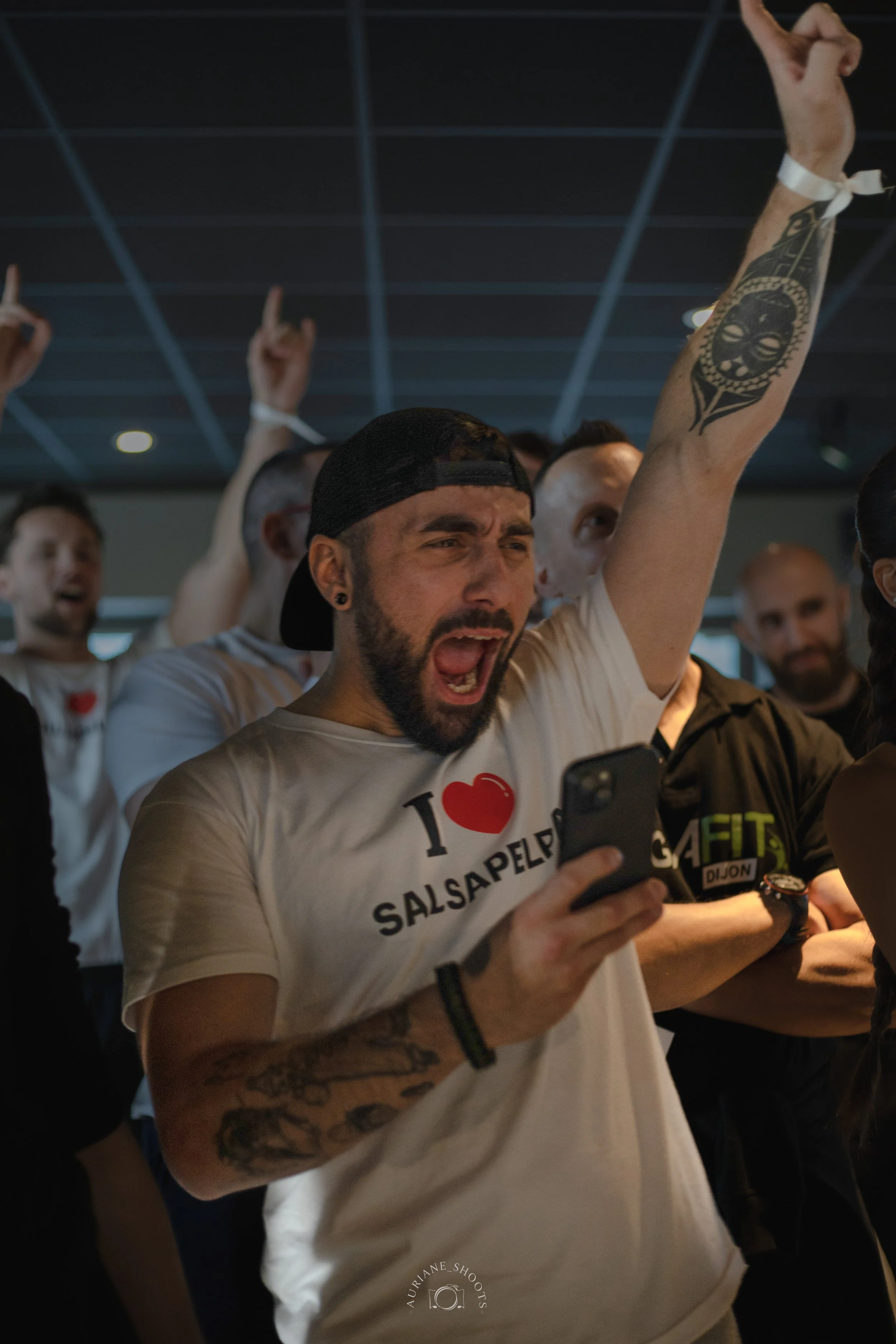Un homme avec une barbe et un tatouage sur le bras, criant tout en regardant son téléphone, entouré de personnes applaudissant et levant les bras, dans une salle.