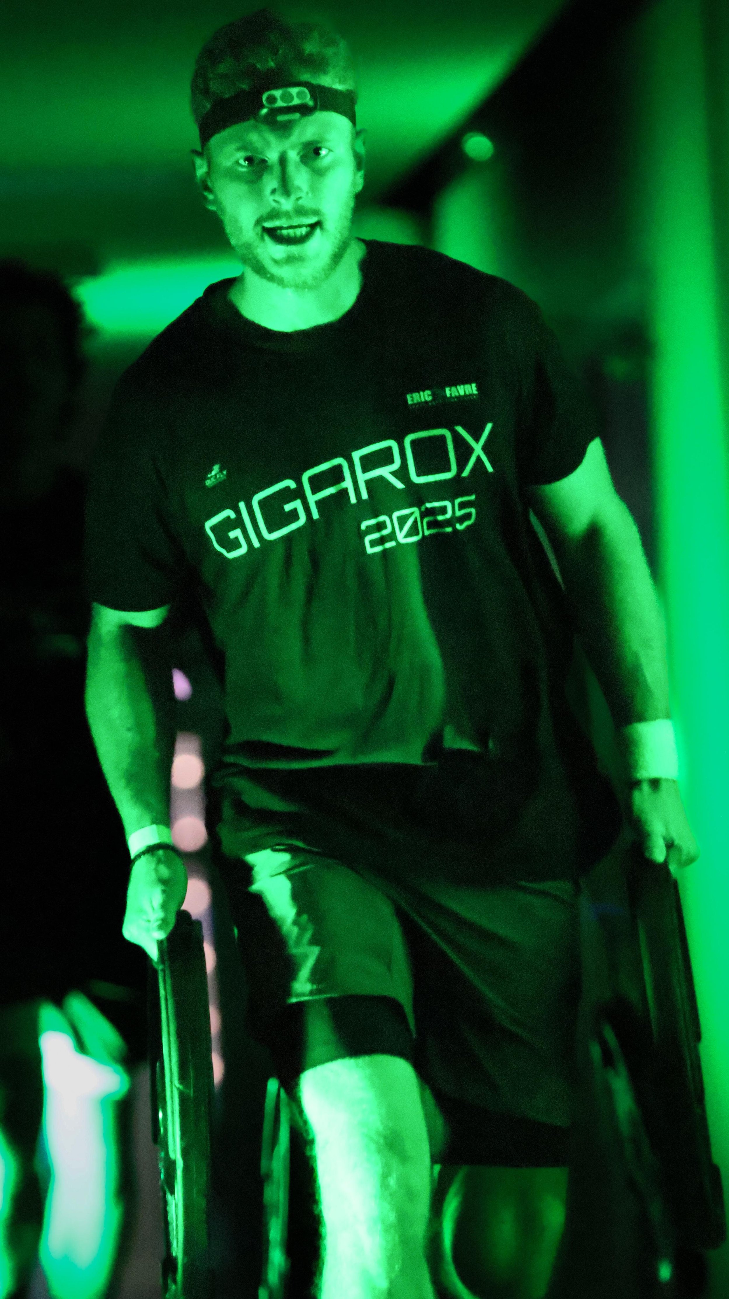 Jeune homme en fauteuil roulant sous un éclairage vert, portant un t-shirt avec le texte 'GIGAROX 2025'