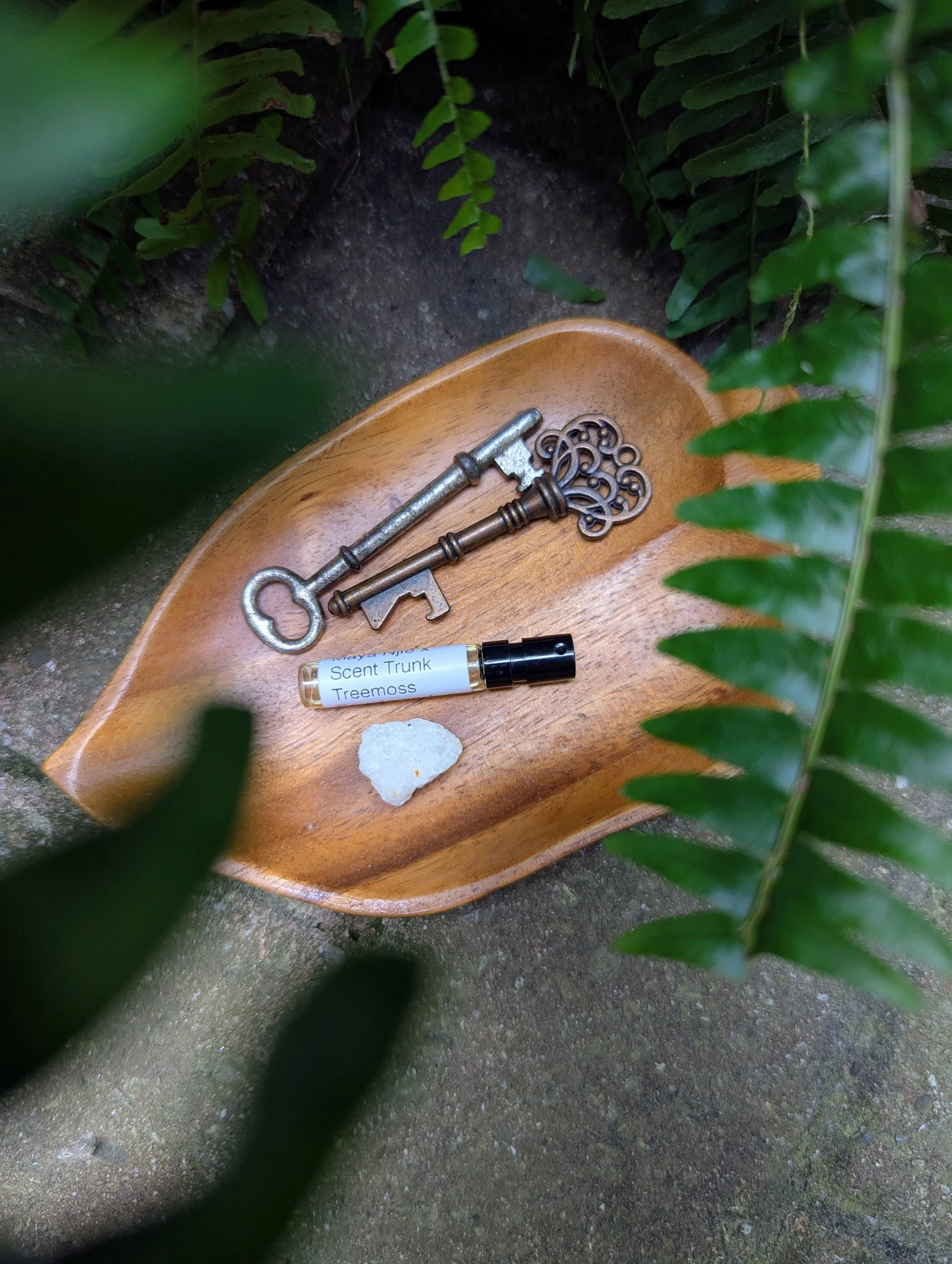 A wooden leaf-shaped dish holding vintage keys, a small perfume bottle labeled 'Scent Trunk Treemoss,' and a white rock, surrounded by green foliage.