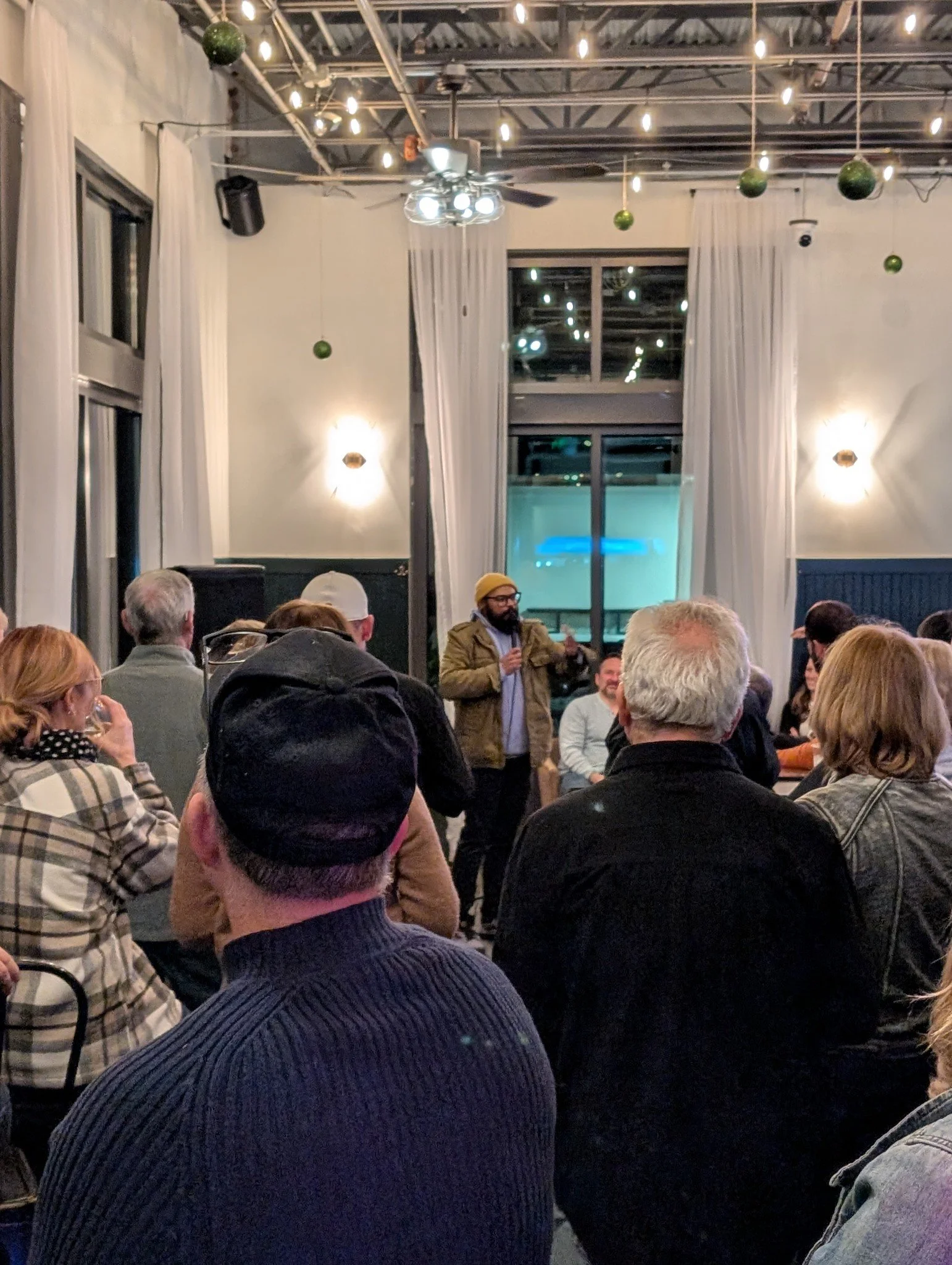 A man with a beard and glasses wearing a yellow beanie and brown jacket speaking into a microphone during a gathering in a decorated indoor venue with audience members seated and white curtains, string lights, and hanging green ornaments.