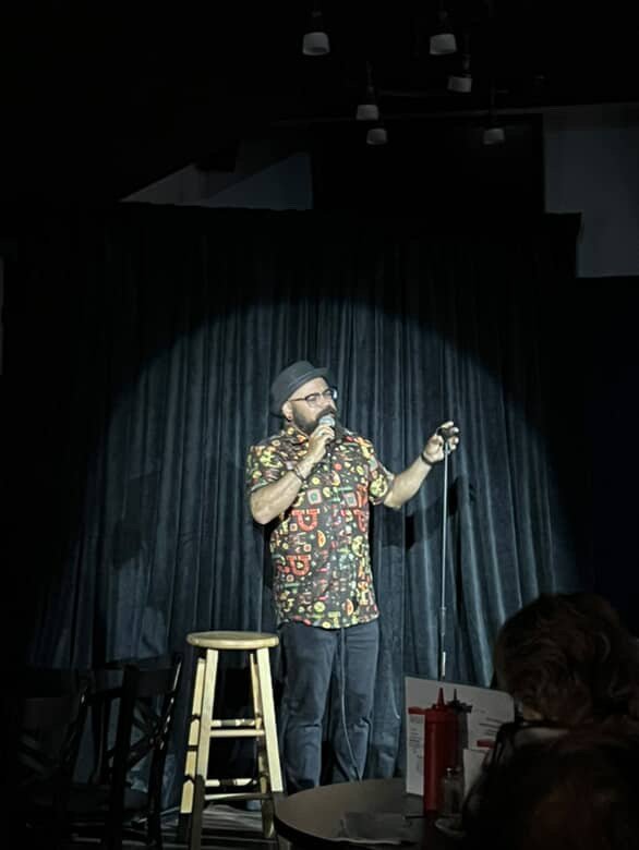 Comedian performing stand-up on stage with a microphone in hand, wearing a patterned shirt, glasses, and a hat, with a small stool beside him and black curtains in the background.