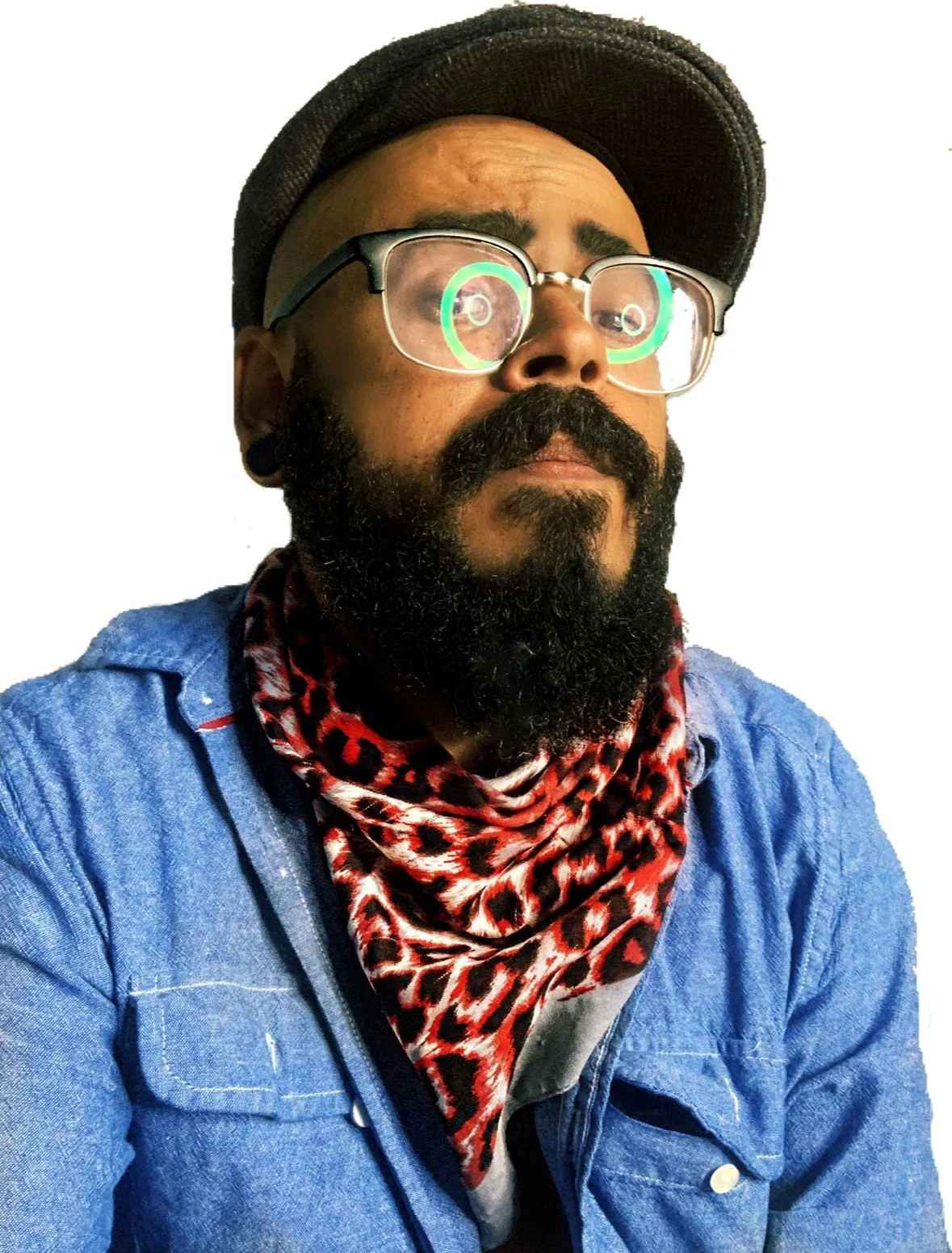 A man with dark hair, beard, and glasses with green reflections, wearing a black cap and a red leopard-print bandana around his neck, dressed in a blue denim jacket.