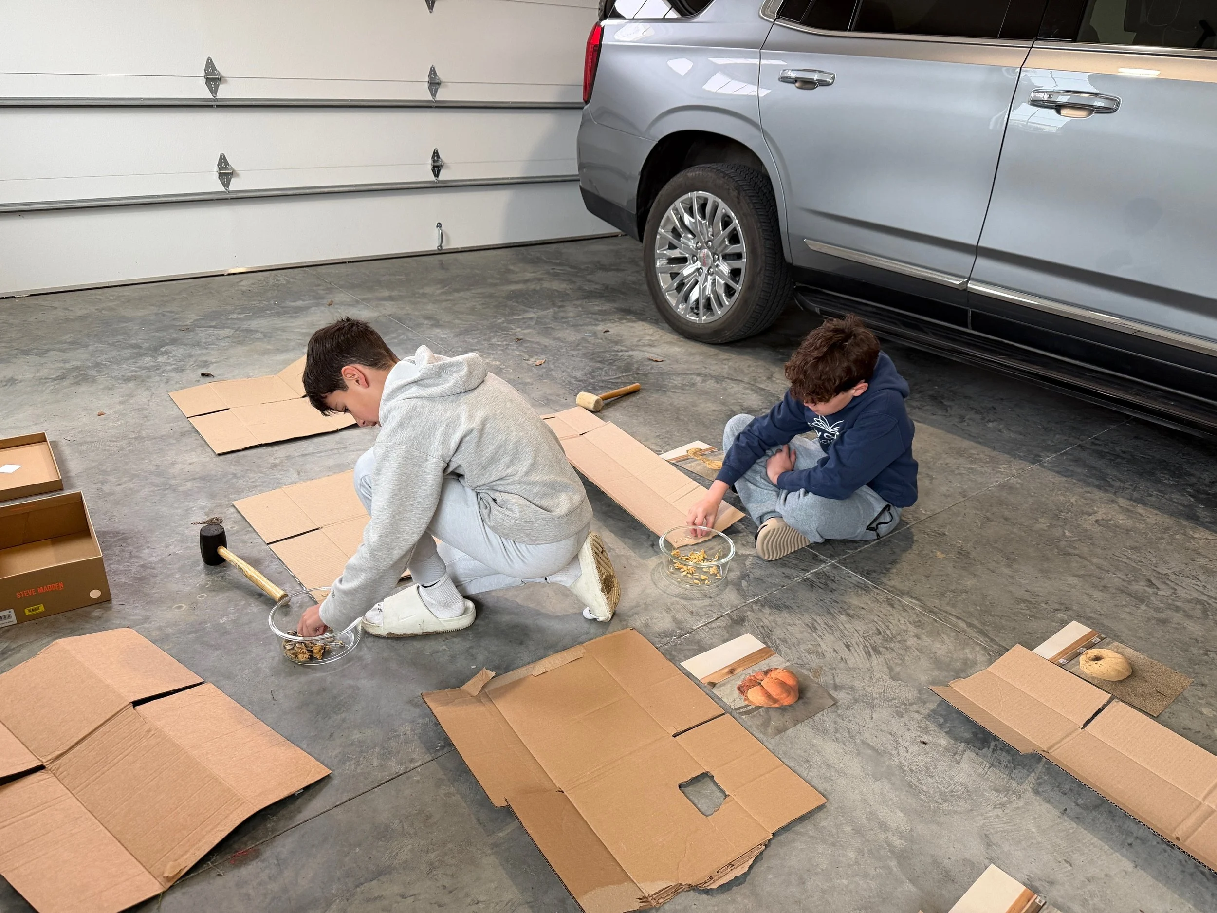 Two boys sitting on a concrete floor assembling cardboard boxes in a garage beside a silver car.
