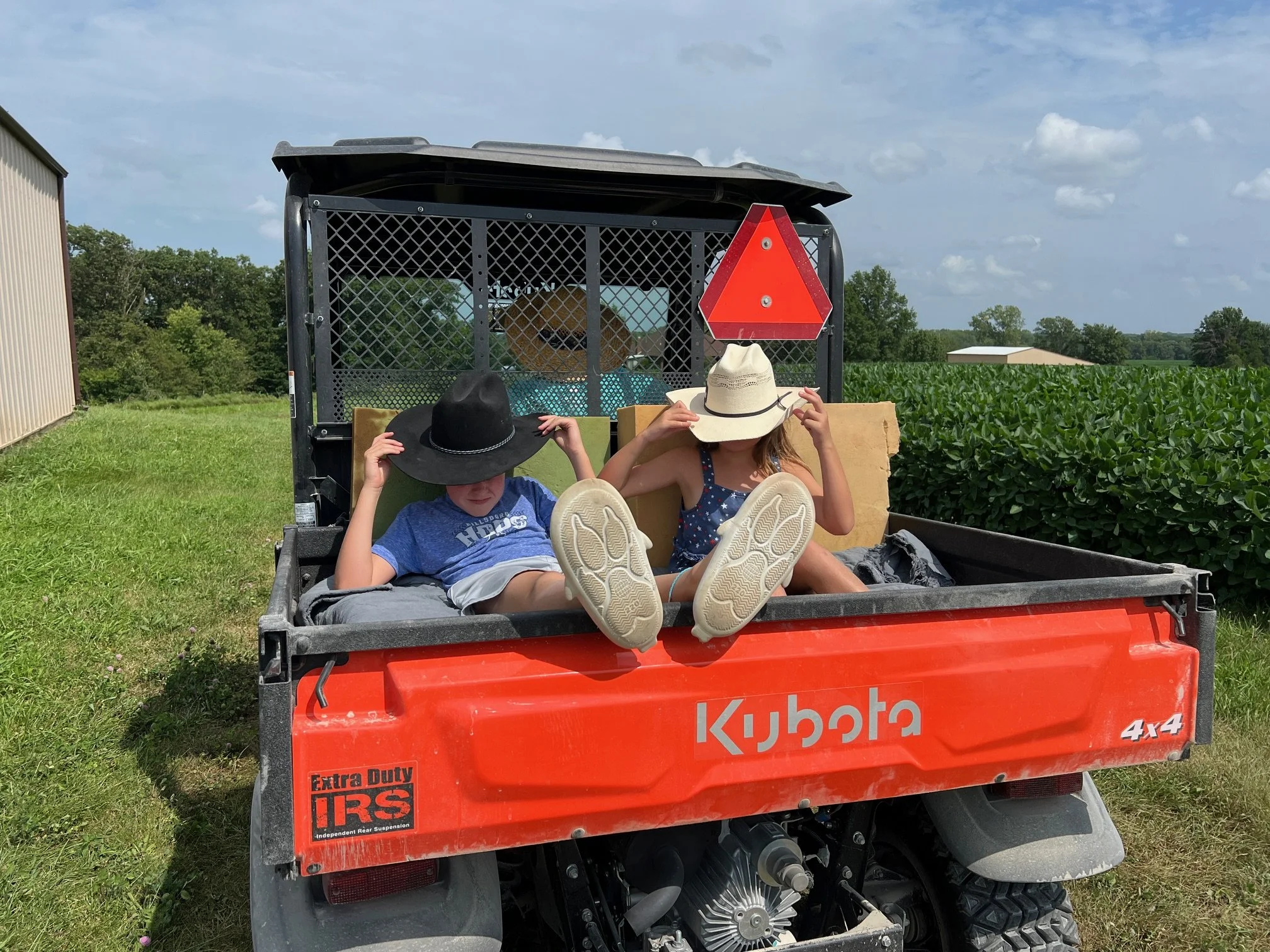 A red utility vehicle with a Kubota logo, two children sitting with their legs out, wearing hats, and a warning reflector in a rural field with farm buildings and green trees in the background.
