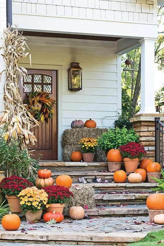 Front porch decorated with pumpkins, mums, hay bales, and fall foliage for autumn.