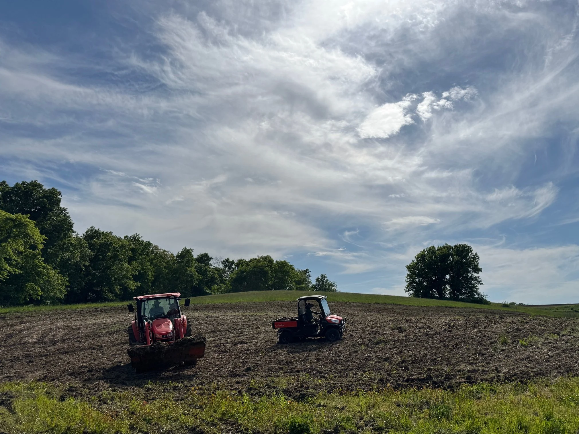 Two red tractors working on a farm field under a partly cloudy sky, with trees in the background.