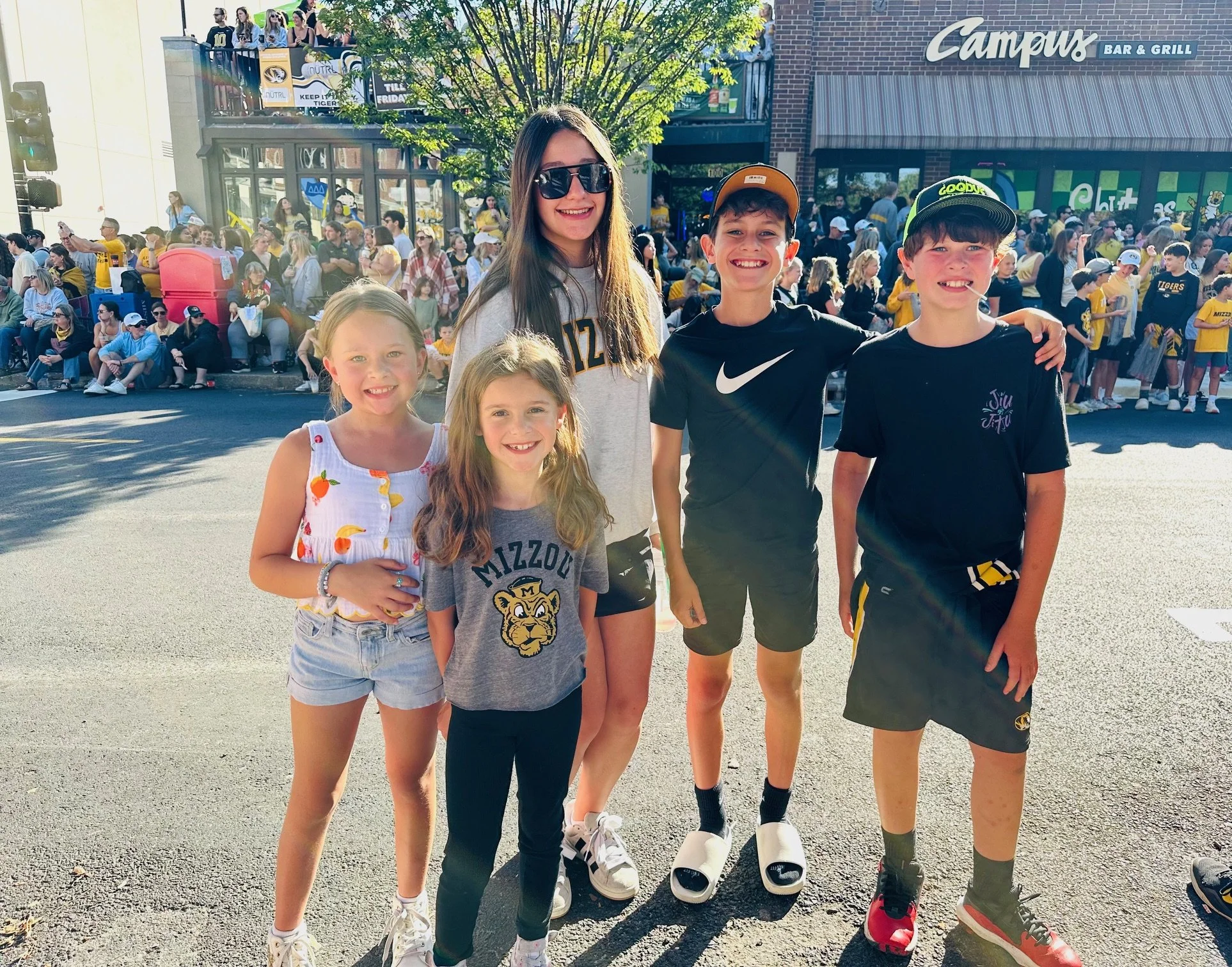 Five children and a young woman standing on a street in front of a large crowd, with some on a balcony, during daytime. The children are smiling, wearing casual summer clothing, and some with hats. The woman has long dark hair, sunglasses, and is wearing a gray shirt. The background features a brick building with a sign for "Campu Bar & Grill" and a tree.